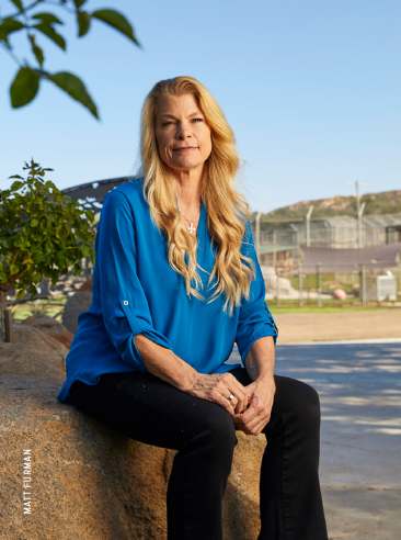 Woman with long blonde hair sitting outdoors on a rock, wearing a blue blouse and black pants, smiling.