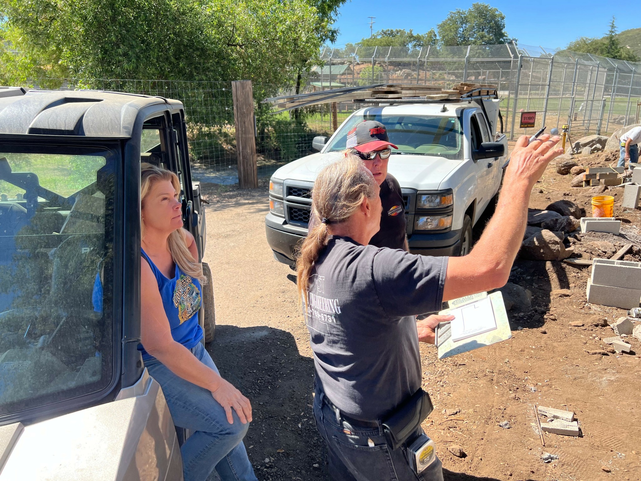 Three people outdoors near a white vehicle, with animals and fencing in the background, one person gesturing with a book.