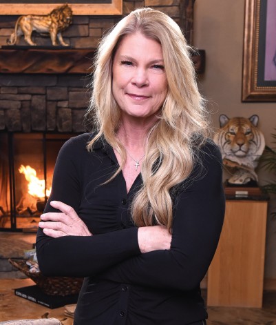 Woman with long blonde hair, arms crossed, smiling in a room with a fireplace and animal decor.