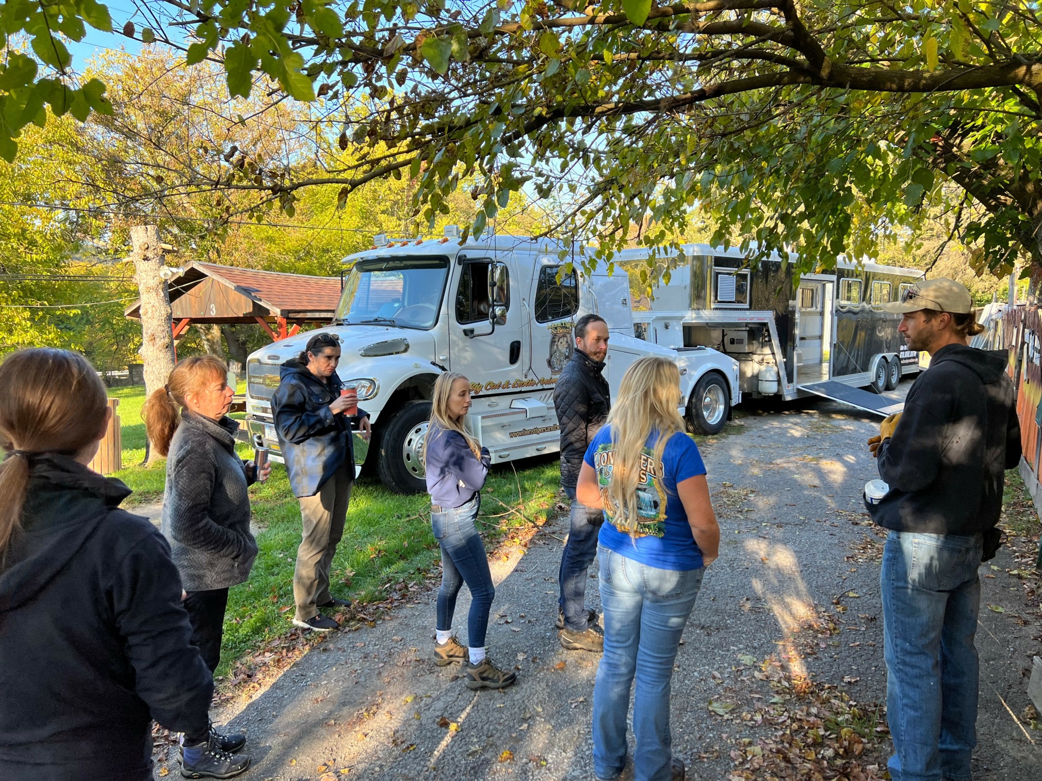 Group of people standing outdoors under trees, listening to a man speaking, with a white vehicle and trailers in background.