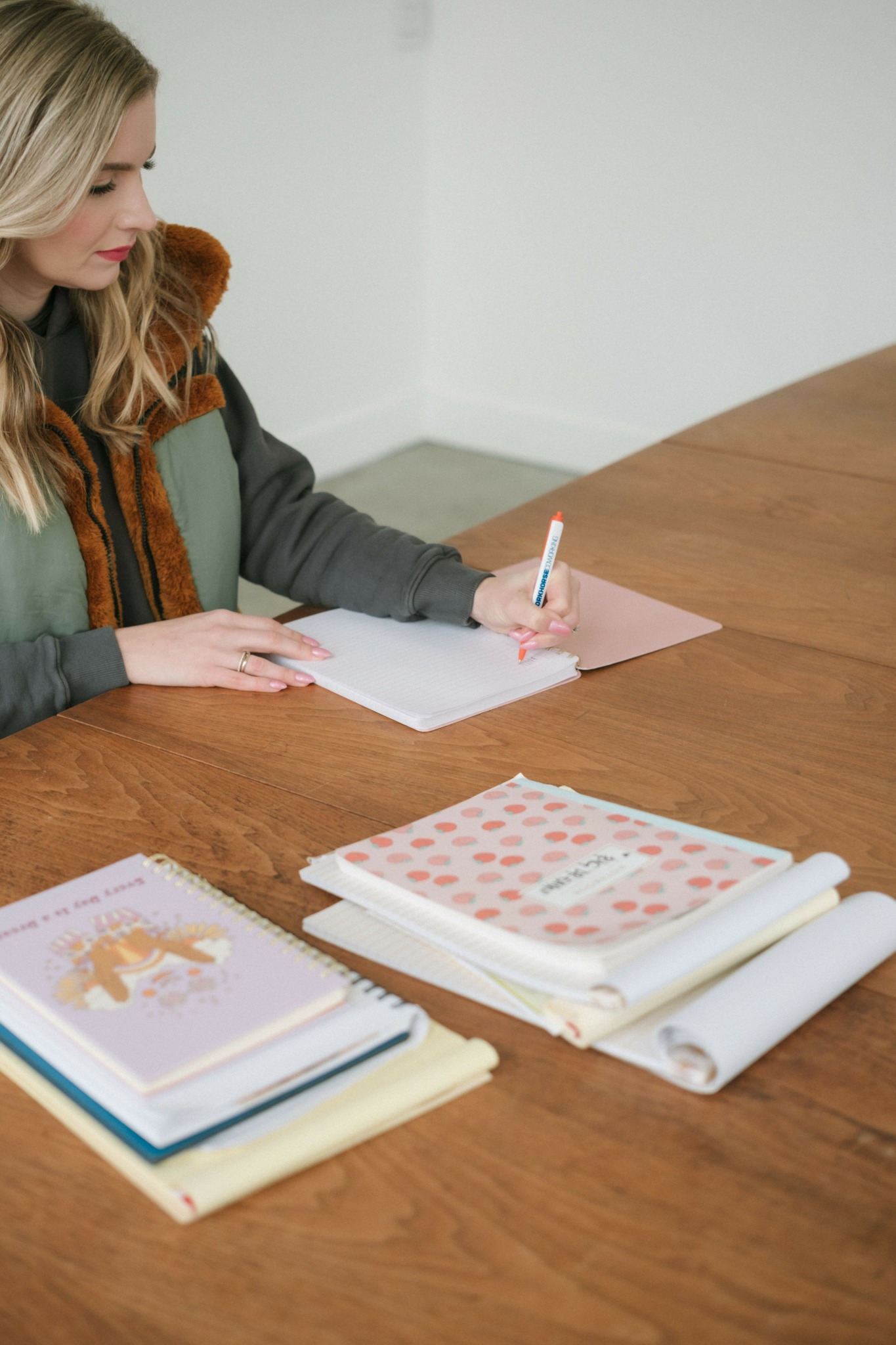 Woman sitting at a wooden table writing on pink paper with notebooks and rolled papers nearby.