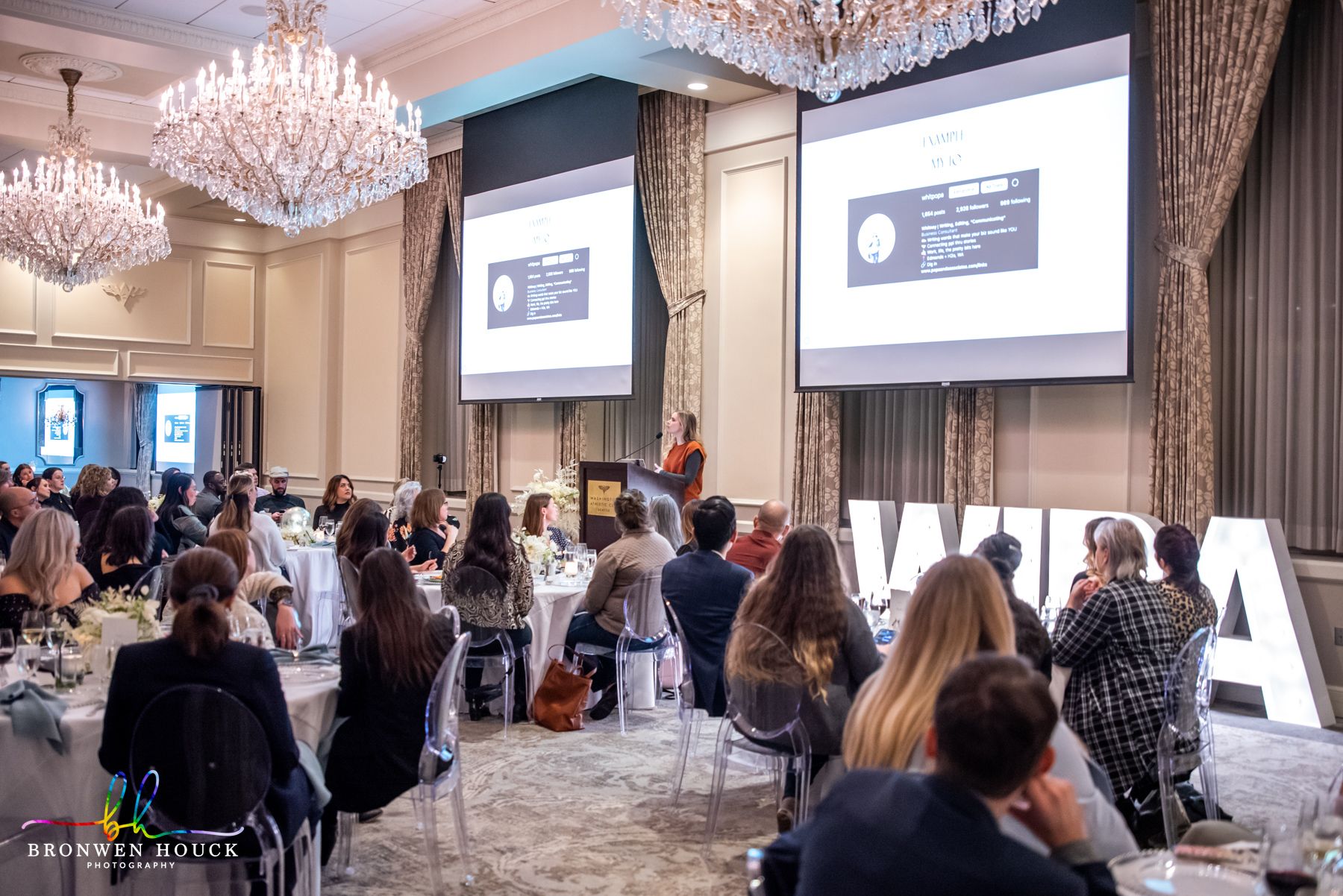 Conference room with audience watching a speaker on stage, large screens display presentation slides, chandeliers hang from ceiling.