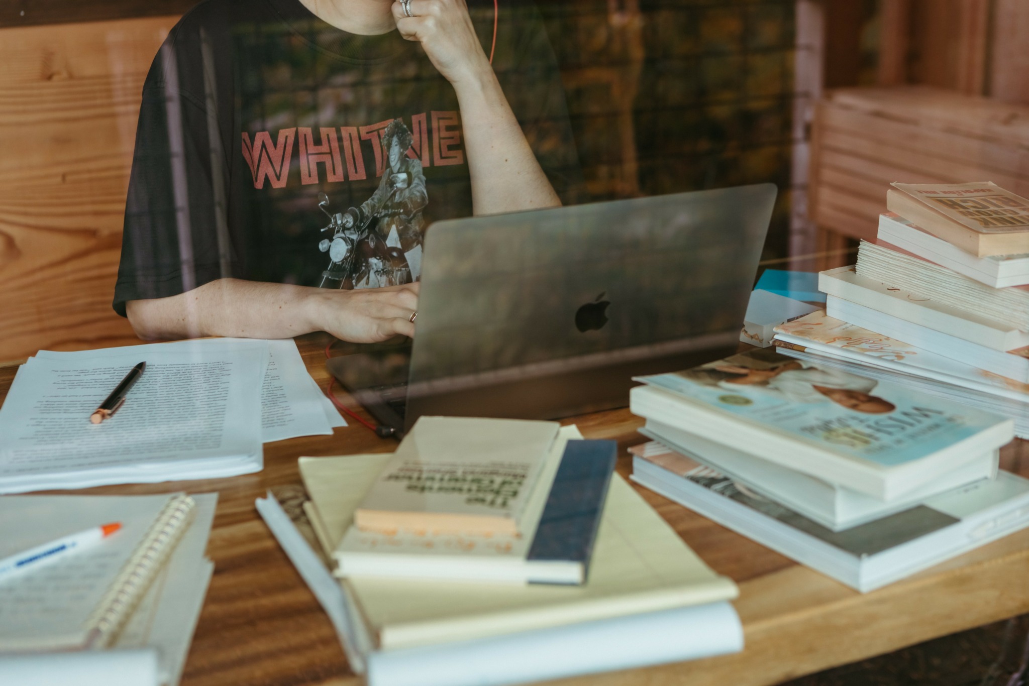 Person sitting at a wooden table with a laptop, books, and papers, wearing a black t-shirt with pink text.