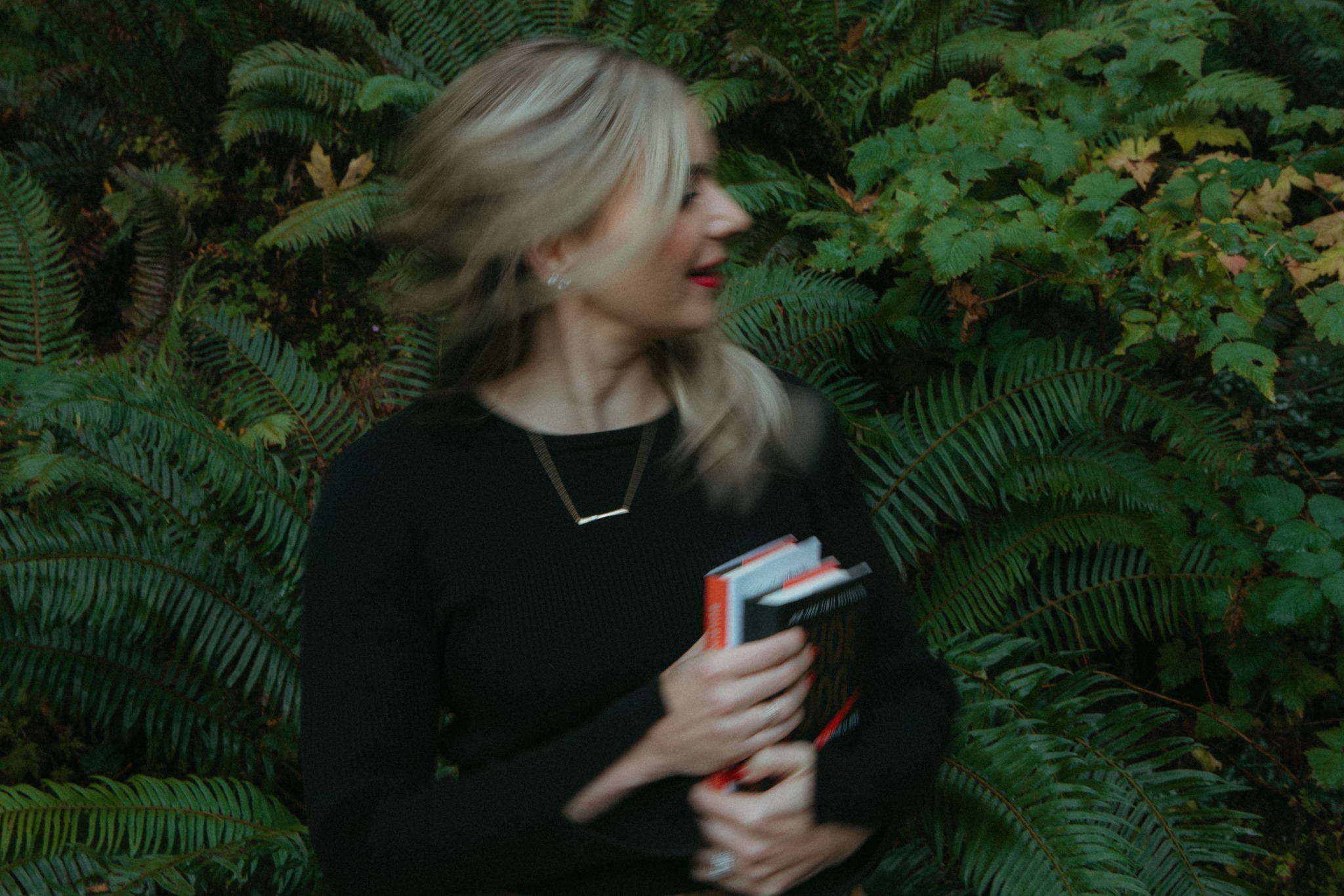 Woman with blonde hair holding books in front of green foliage background.