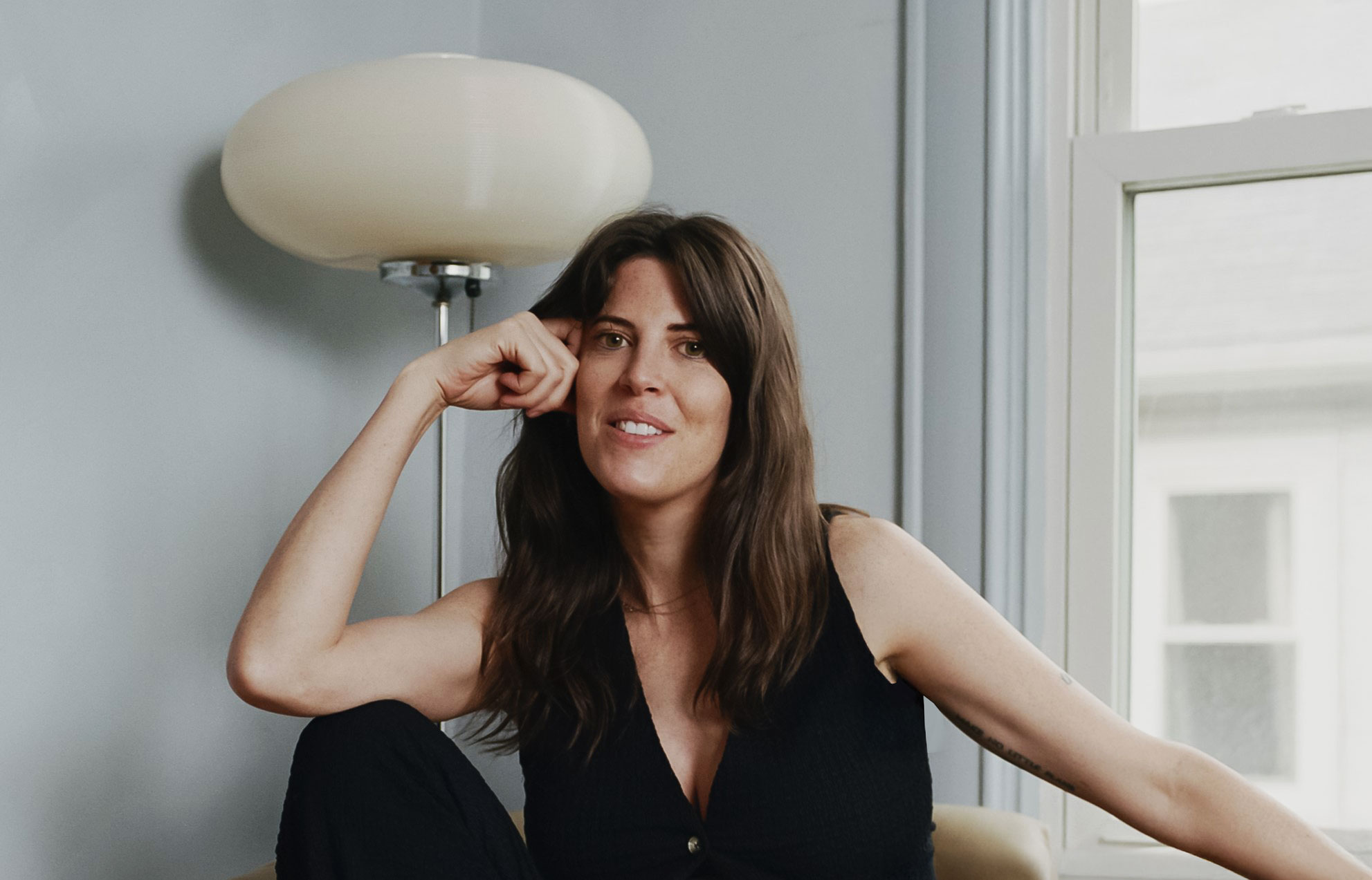 Woman with dark hair smiling, sitting near a window with a lamp behind her.