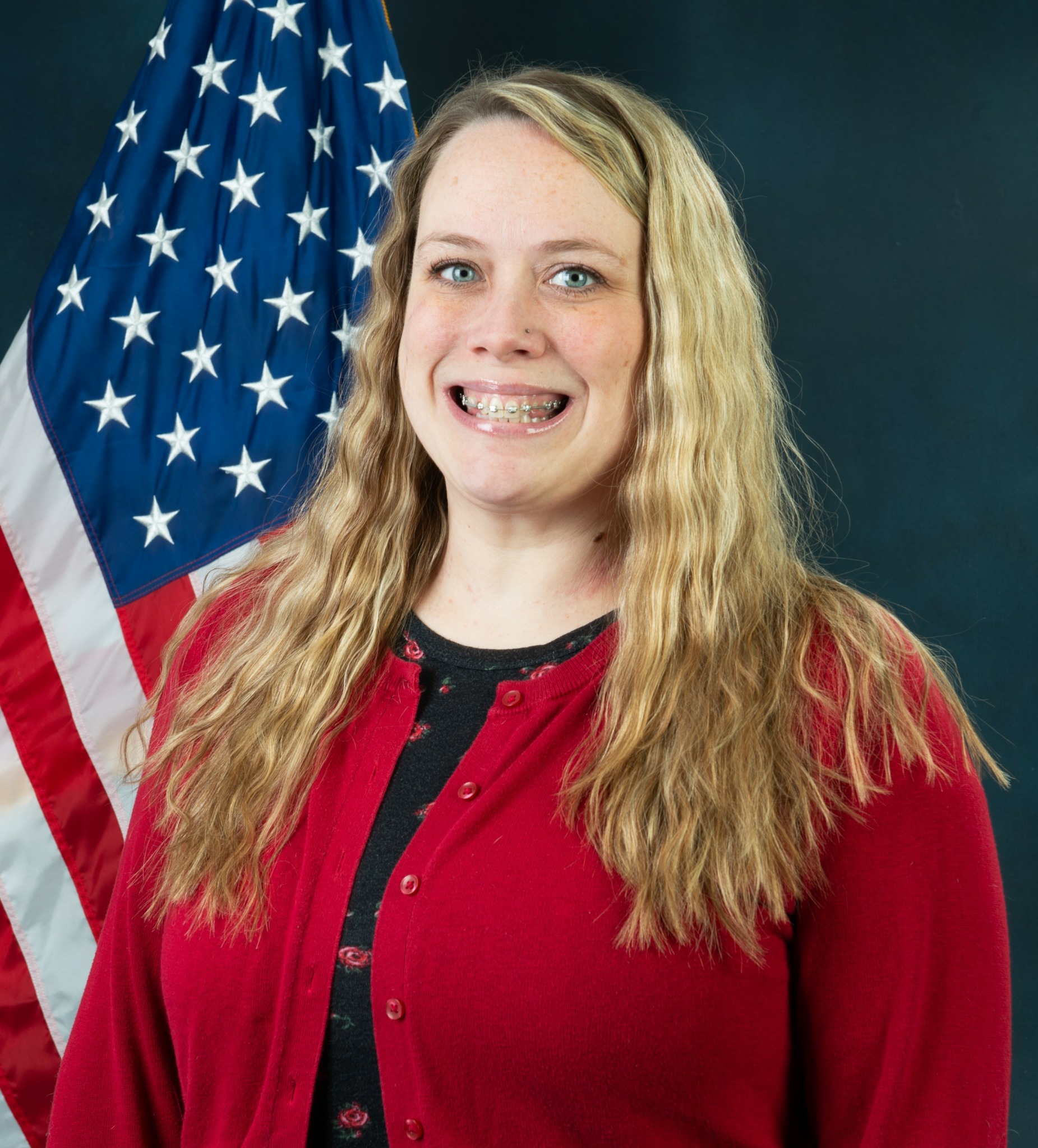 Woman with long wavy blonde hair smiling, wearing a red cardigan, standing in front of an American flag.