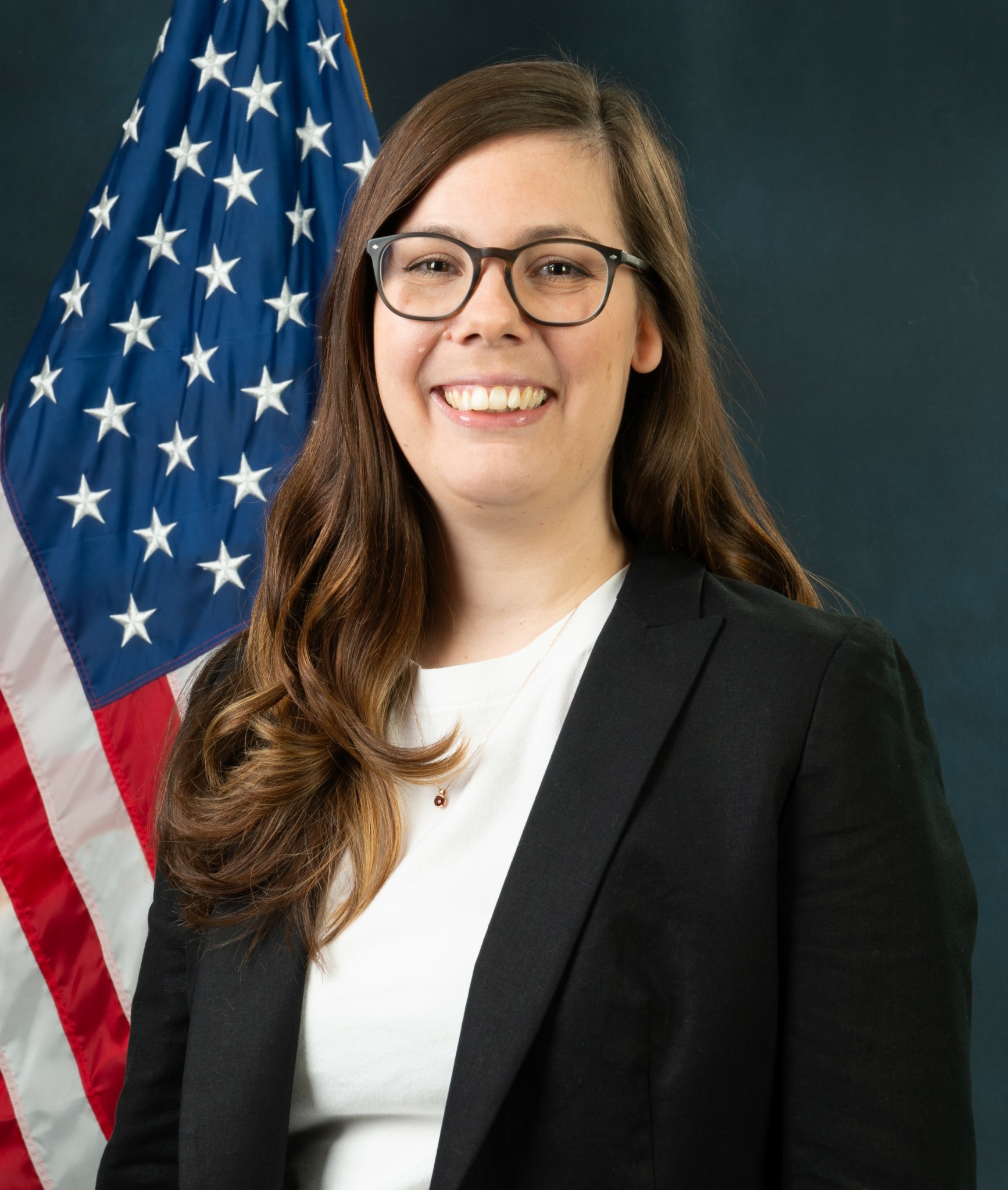 Woman with long brown hair, glasses, smiling, wearing a black blazer and white top, standing in front of an American flag.