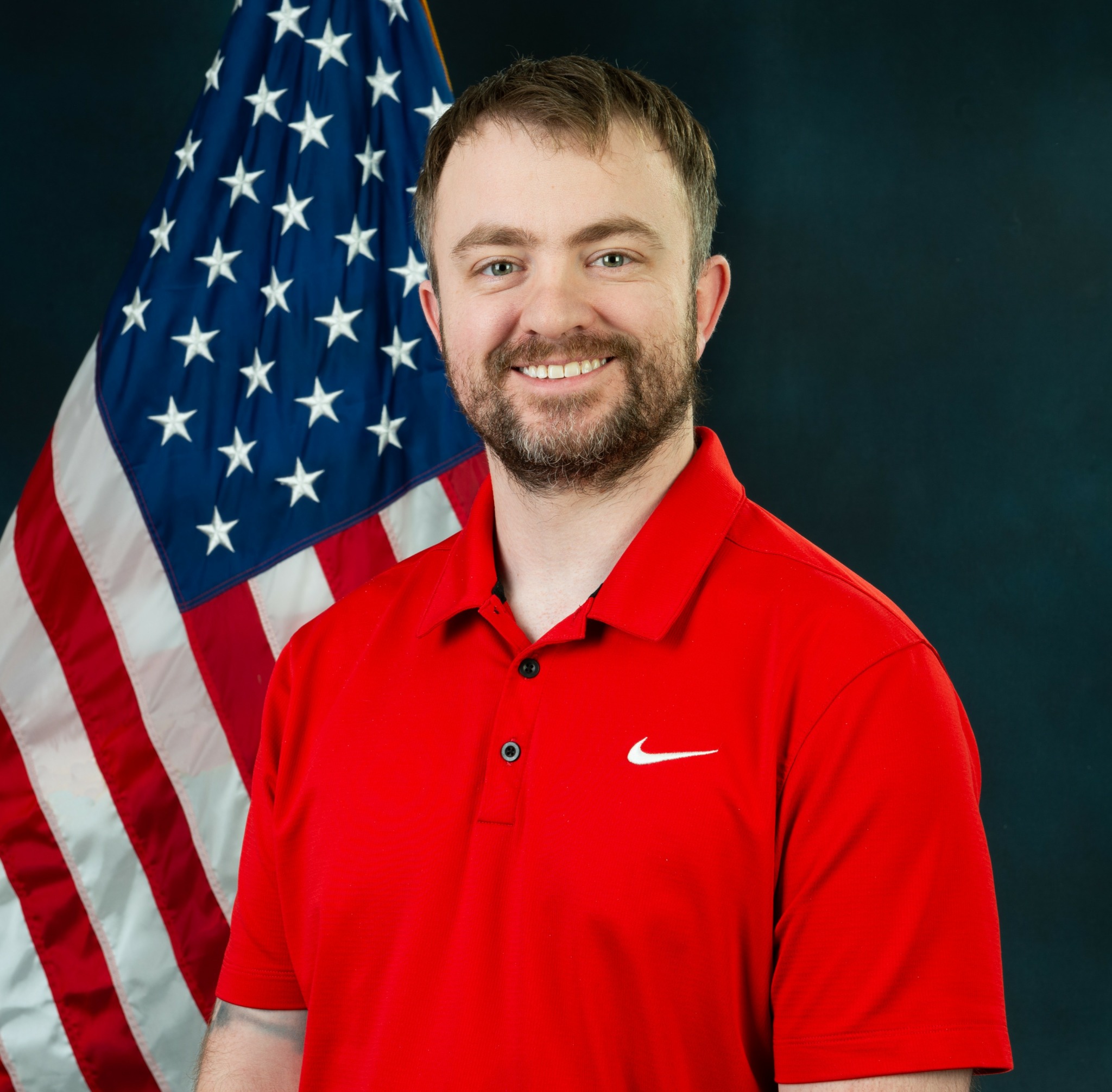 Man with beard in red Nike polo shirt standing in front of American flag, smiling.