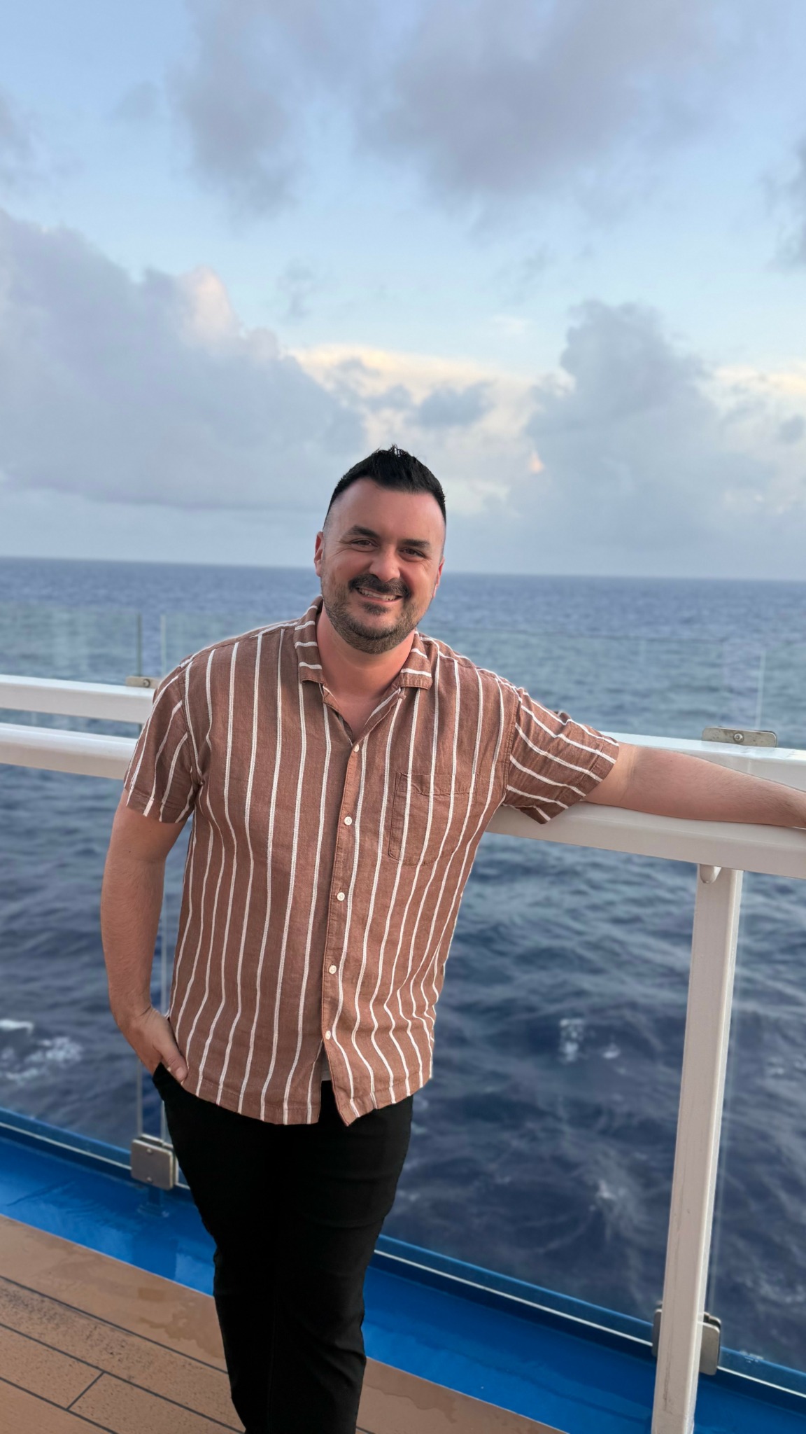 Man with short dark hair and beard smiling, standing on a ship deck with ocean and sky in background.