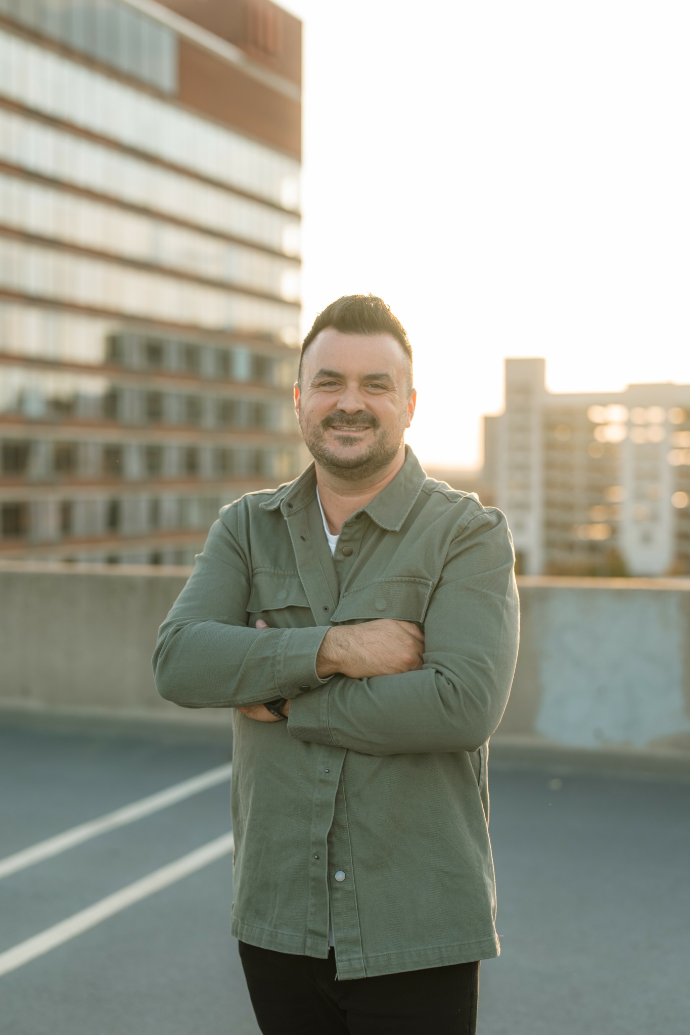 Man with short dark hair and beard standing outdoors with arms crossed, city buildings in background.