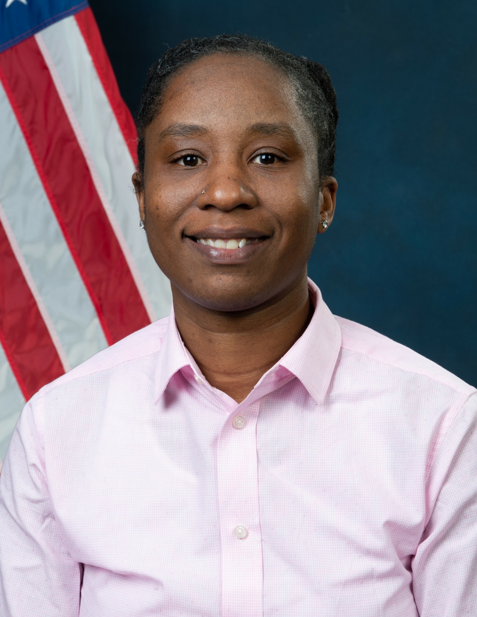 Woman with short hair wearing a light pink collared shirt, smiling, with an American flag in the background.