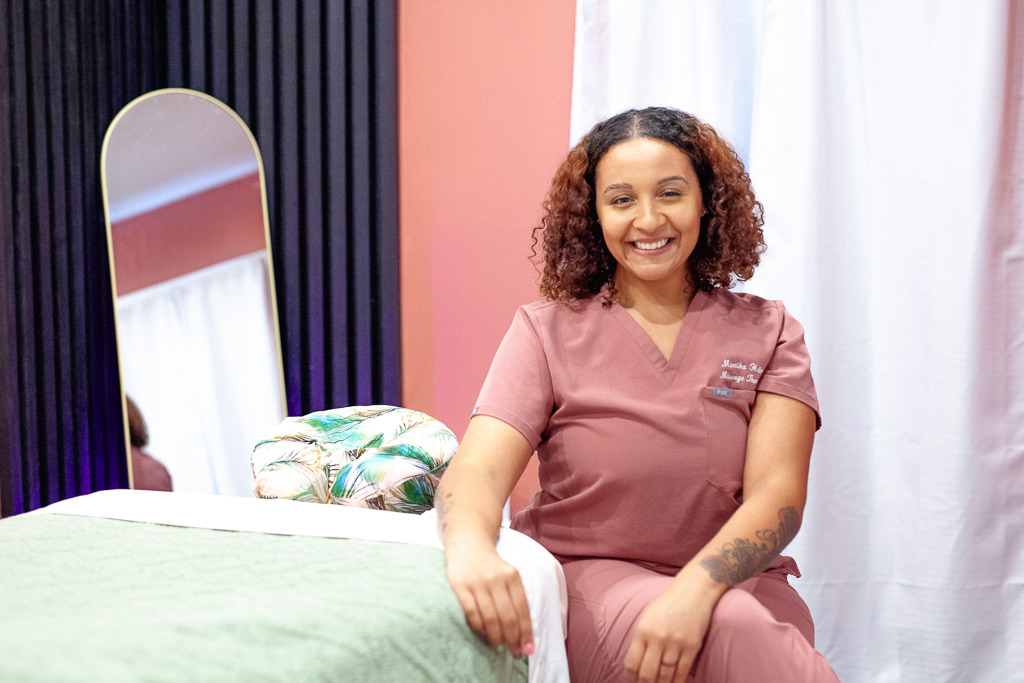 Smiling woman in pink scrubs sitting beside a bed with colorful pillows in a room with pink and white walls.