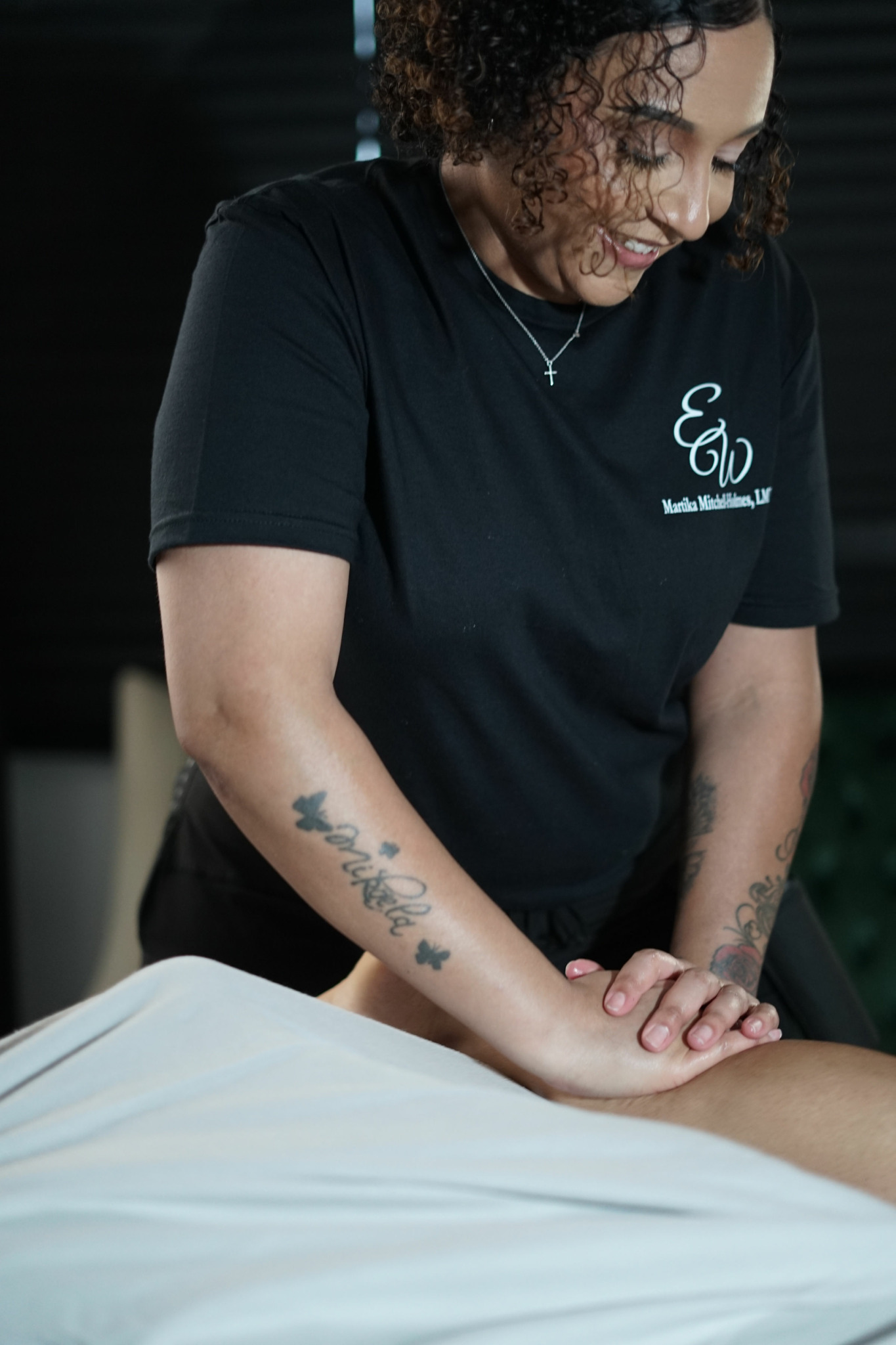 Woman with curly hair smiling, wearing a black T-shirt, leaning over a bed or table, tattooed arms visible.