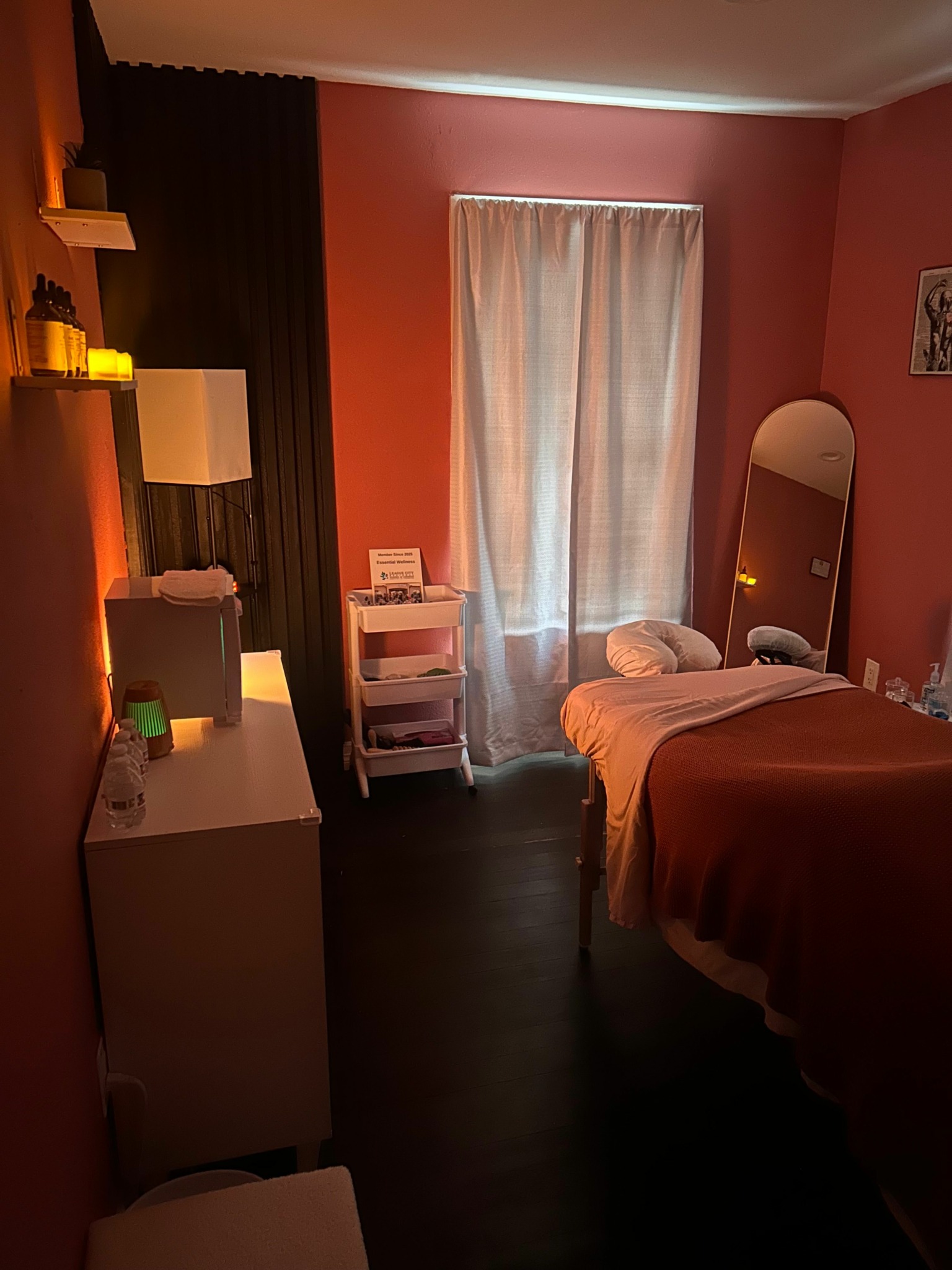 Hotel room with bed, window with curtains, and a small table with items, illuminated by soft lighting.