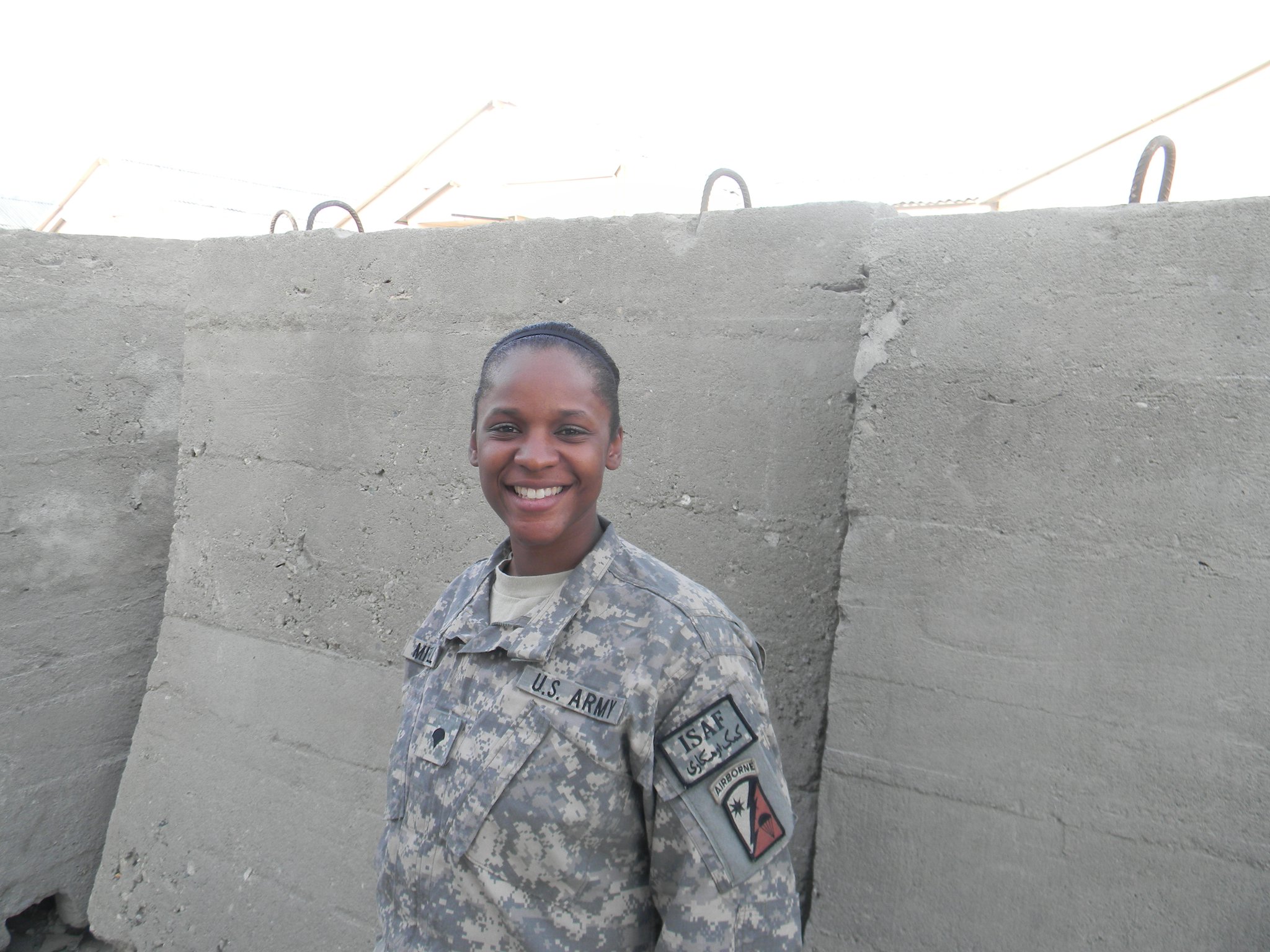 Smiling person in military uniform standing outdoors against a concrete wall.