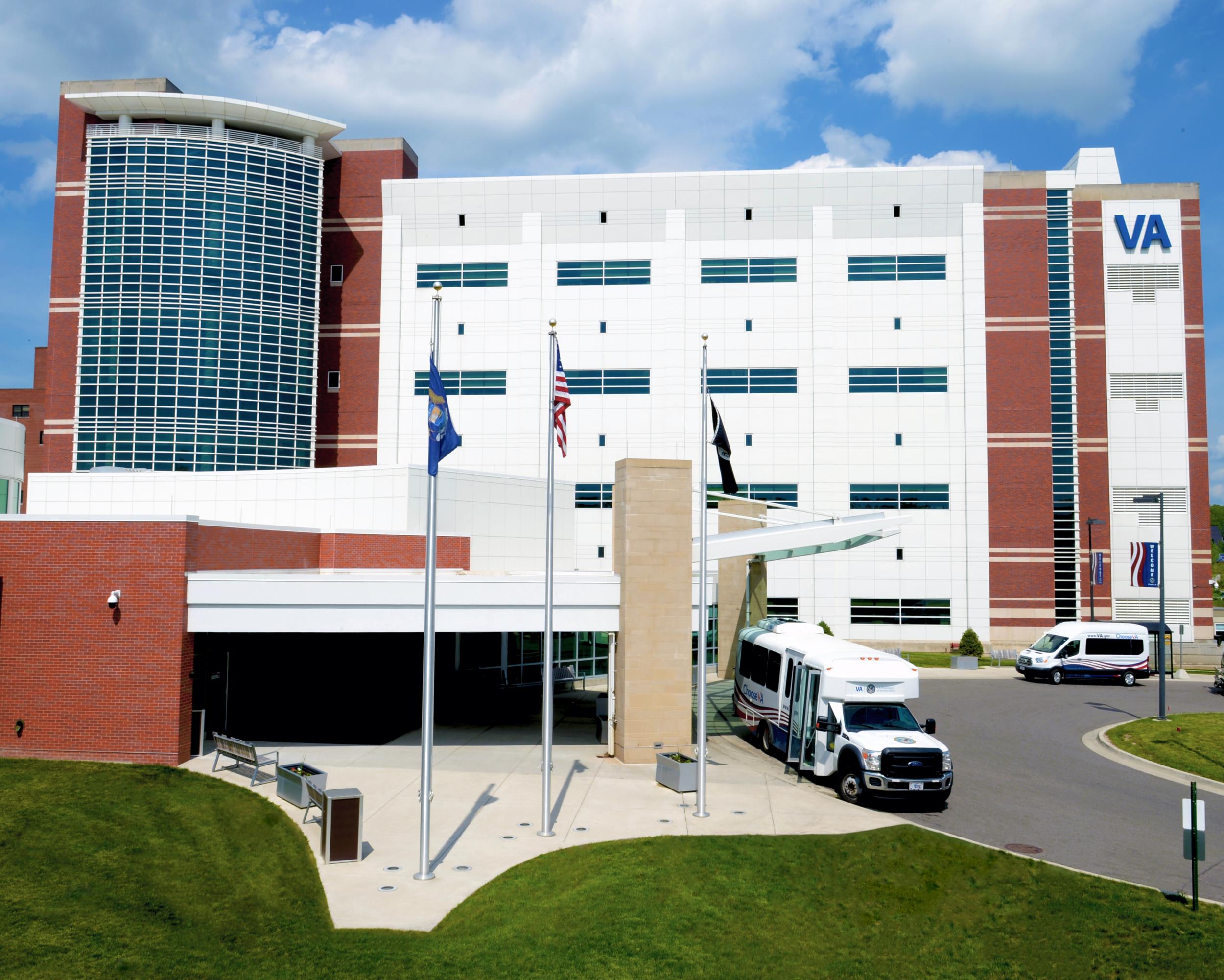 Hospital building with flags, bus, and cars outside, VA logo visible on the right side, blue sky with clouds.