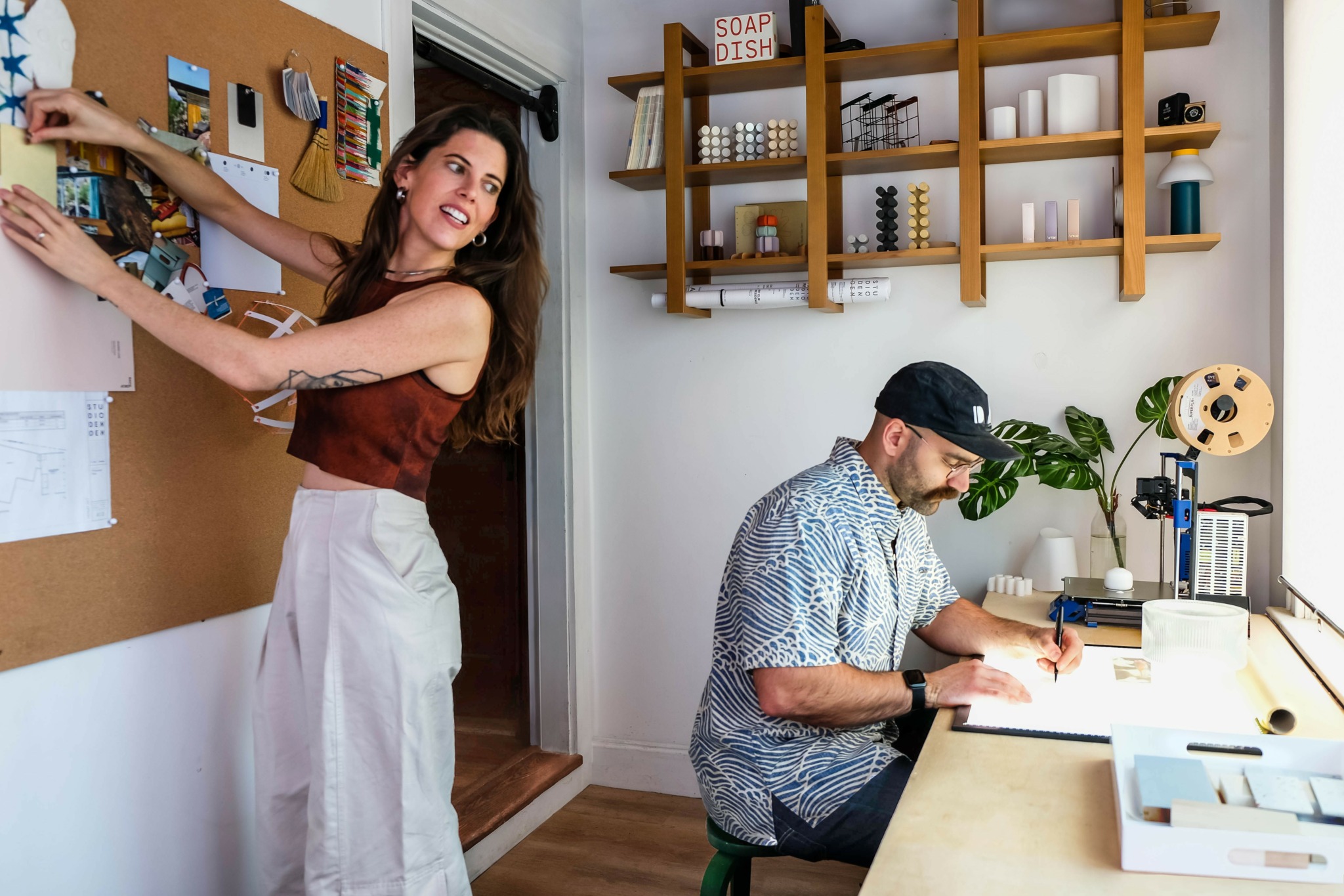 Woman pinning notes on corkboard, man working at desk with plants and office supplies, shelves with books and decor in background.