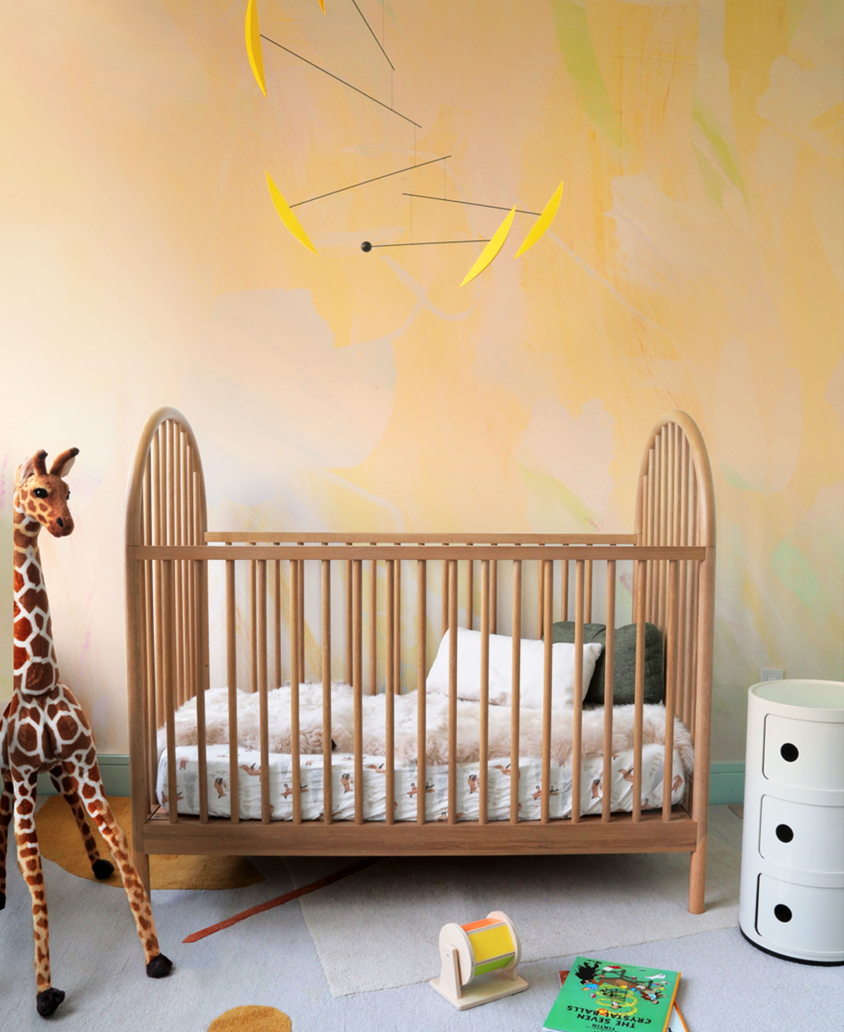 Baby crib with bedding, a giraffe plush toy, a small colorful toy, and a white storage unit in a nursery.