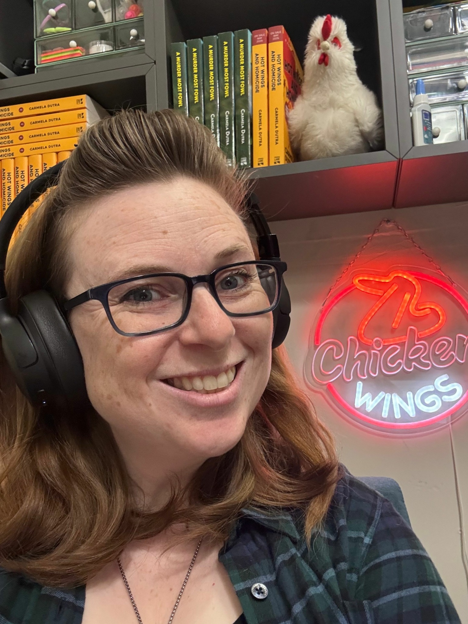 Woman with glasses and headphones smiling, bookshelf with books and a stuffed animal in the background, neon sign, and plaid shirt.