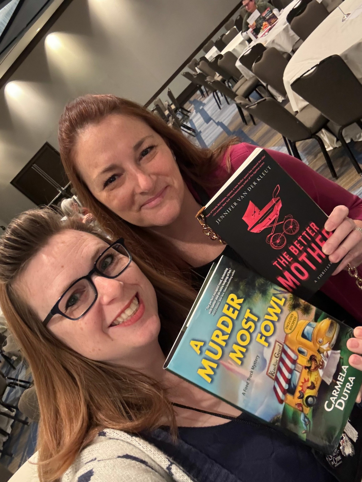 Two women lying down with books and a magazine, smiling at the camera, in an indoor setting.