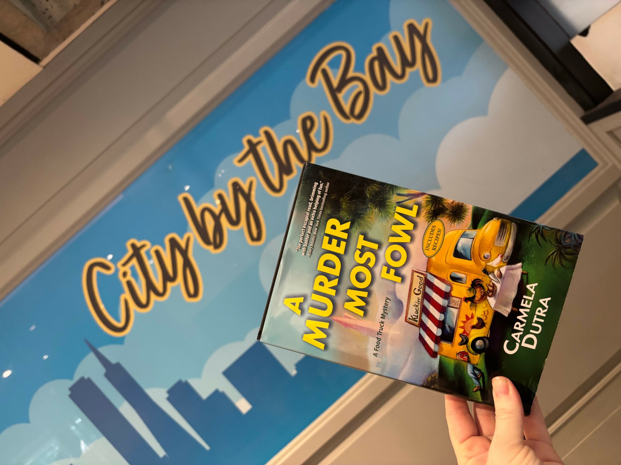 Person holding a book titled 'A Murder Most Fowl' in front of a sign that says 'City by the Bay'.
