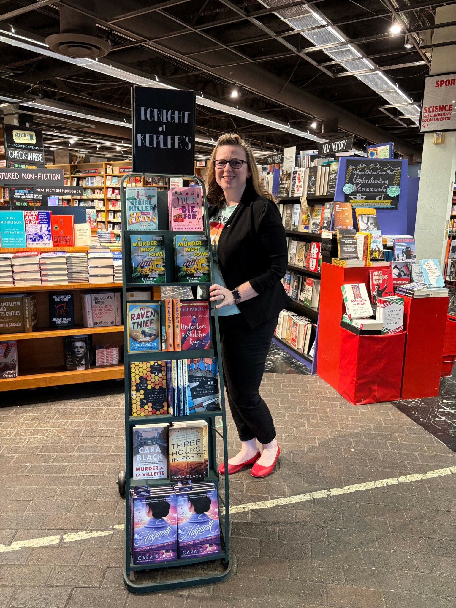 Woman standing in bookstore aisle, surrounded by shelves of books, with a display table nearby.