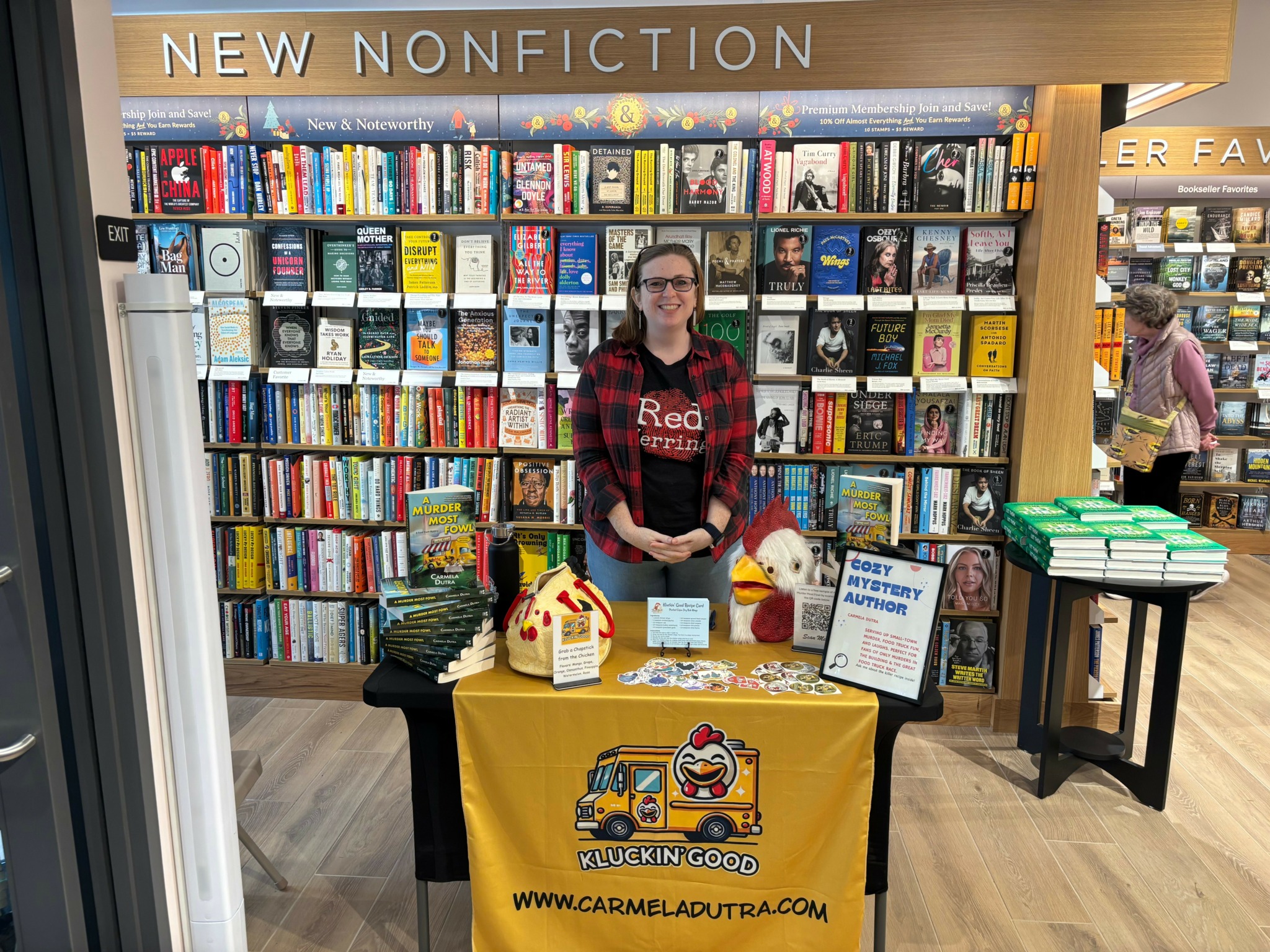 Person standing behind a table with a yellow cloth in a bookstore, bookshelves in background.