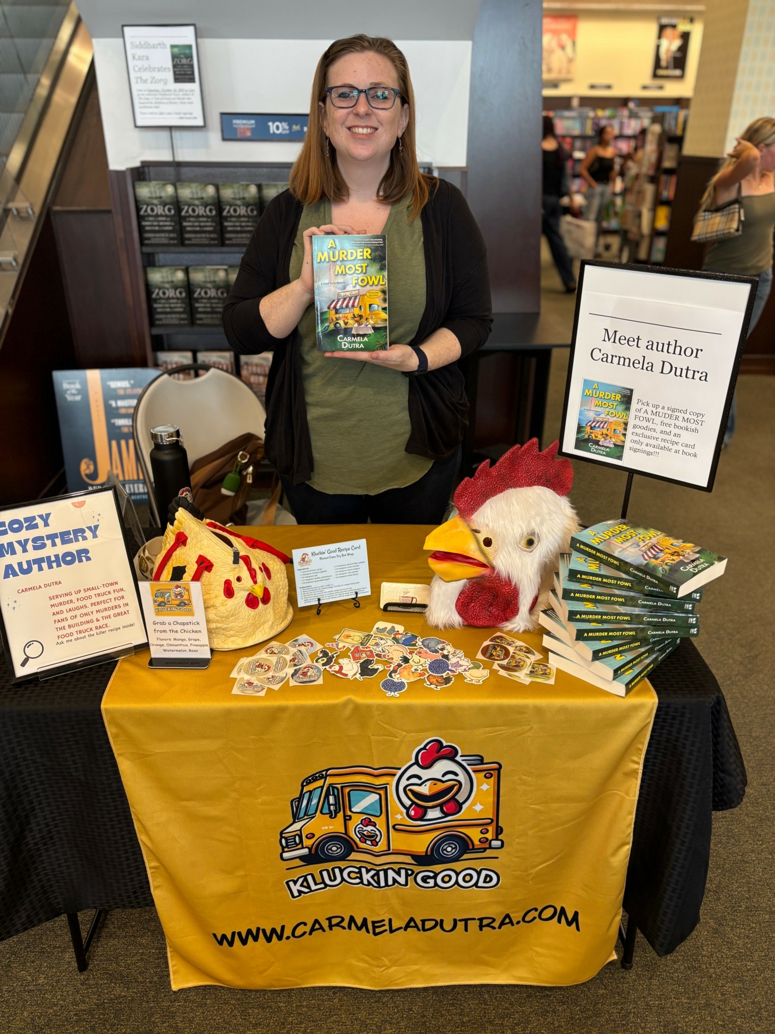 Woman holding a book at a table with books, plush toys, and promotional materials in a bookstore or event space.