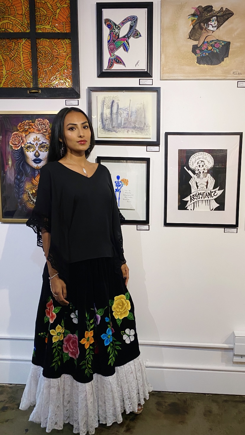 Woman standing in front of wall with framed artwork, wearing a black top and a floral skirt with white lace hem.