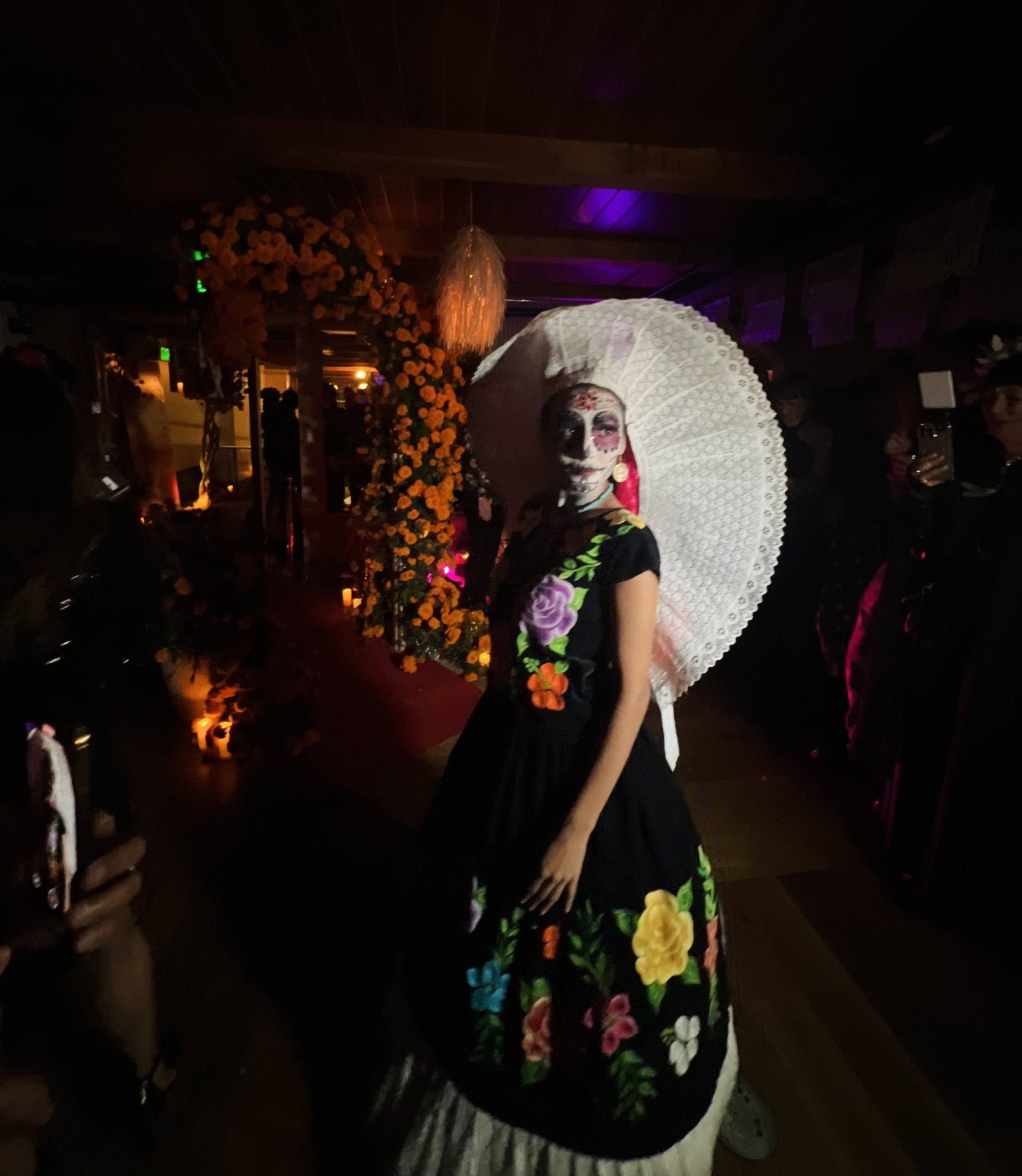 Person dressed as a skeleton with colorful floral dress, face paint, holding a white parasol, in a decorated indoor setting.
