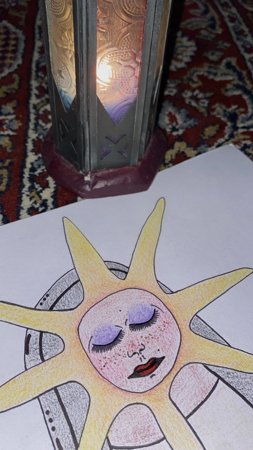Decorative lantern with a lit candle and a drawing of a sun with a face on paper nearby.
