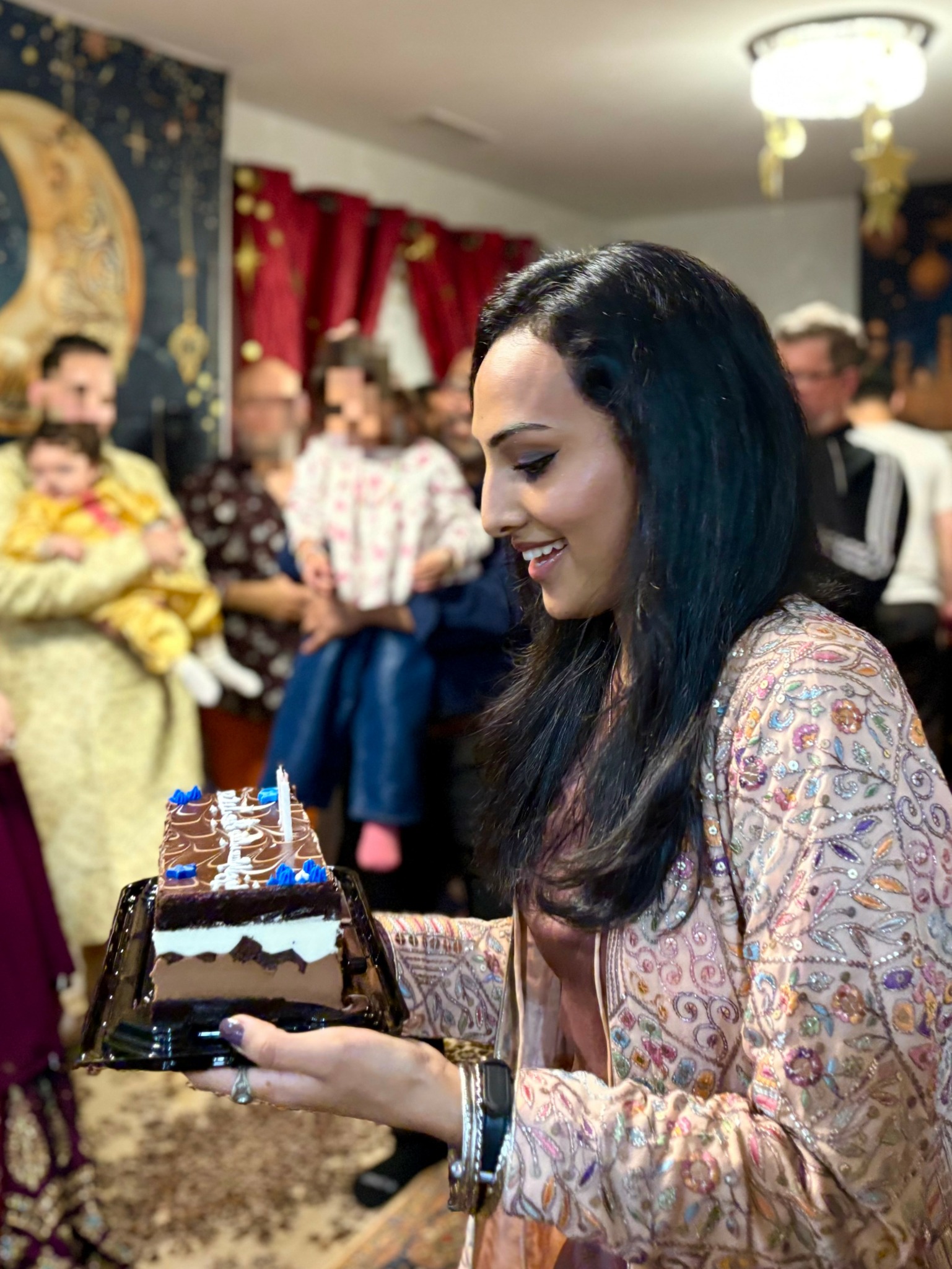 Woman holding a birthday cake with blue candles, smiling at a celebration with people in the background.