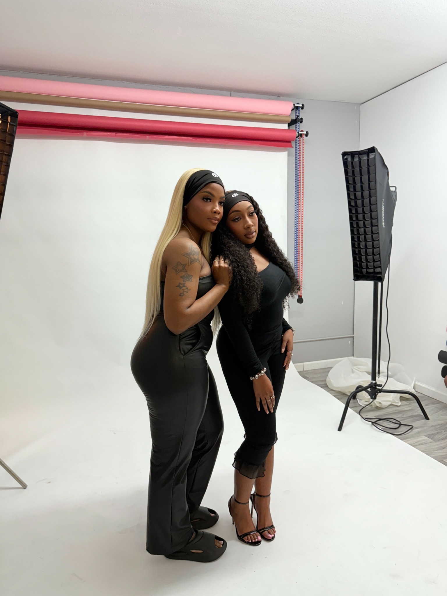 Two women stand in front of a white backdrop, one in black pants and top, the other in a black dress and heels.