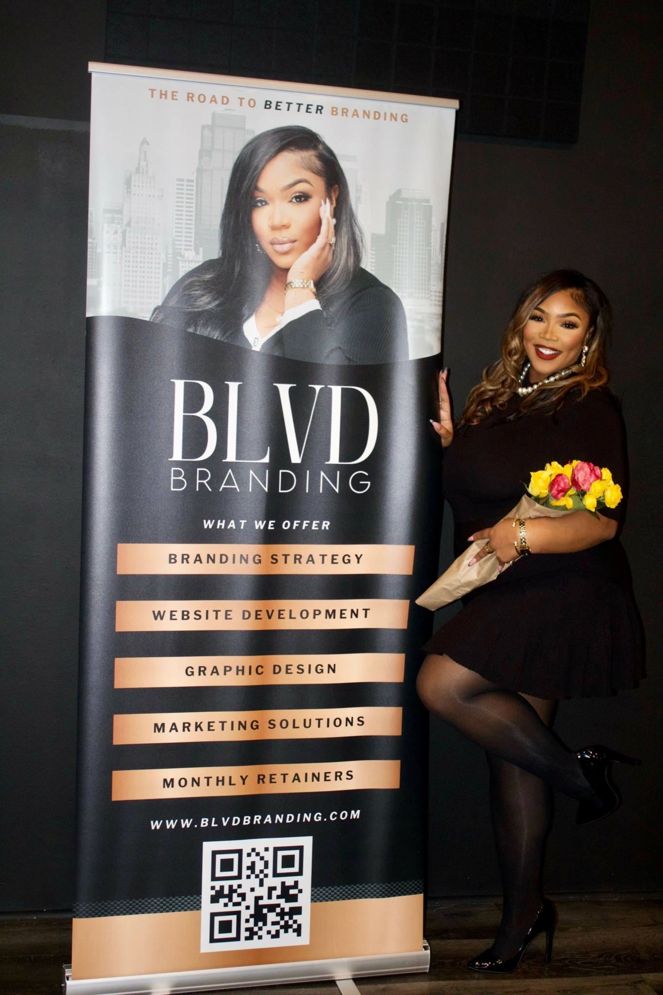 Woman in black dress holding a yellow bouquet, standing next to a BLVD Branding banner with a woman's face and text.