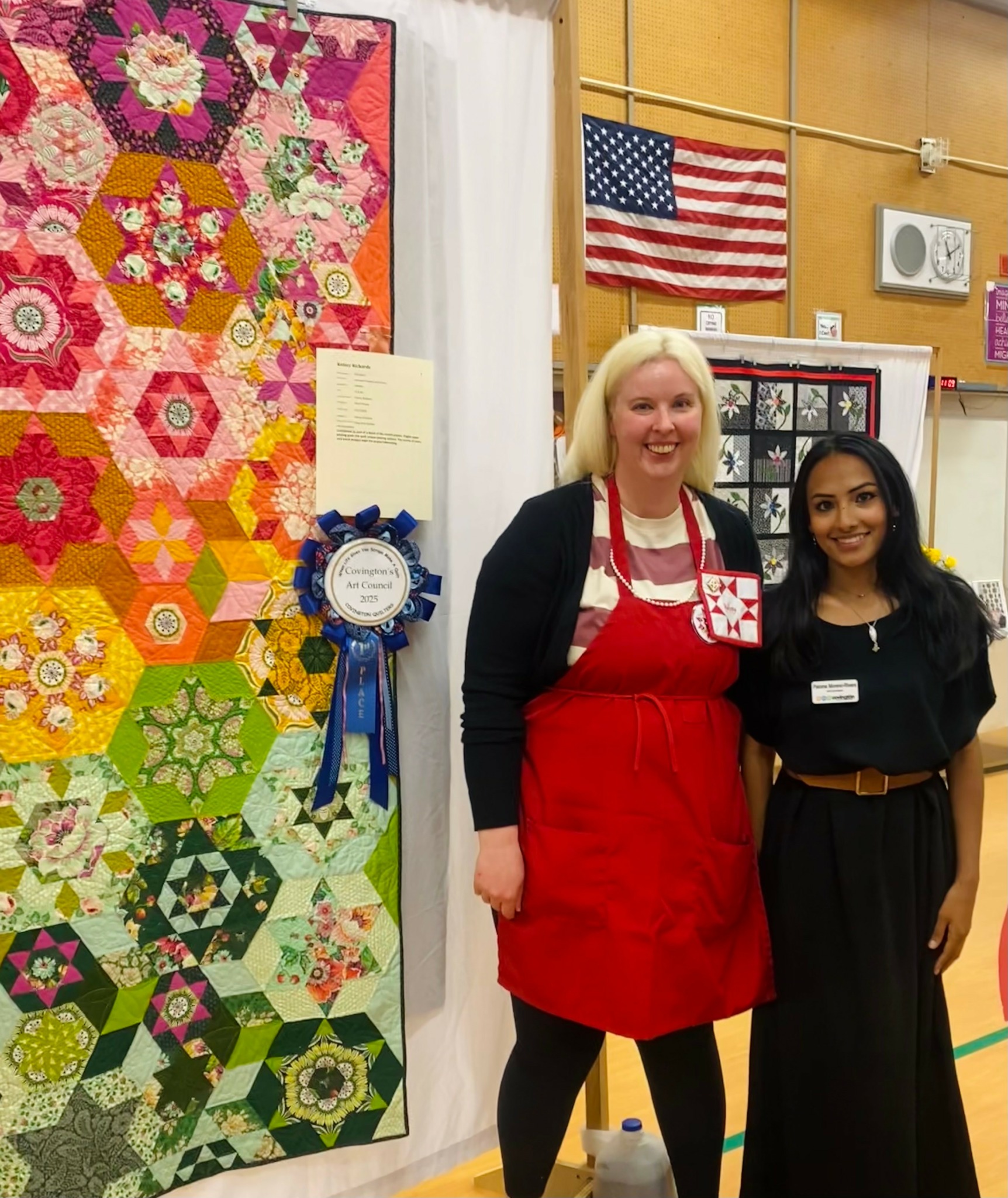 Two women standing next to a colorful quilt display, smiling, in a room with an American flag and bulletin board.