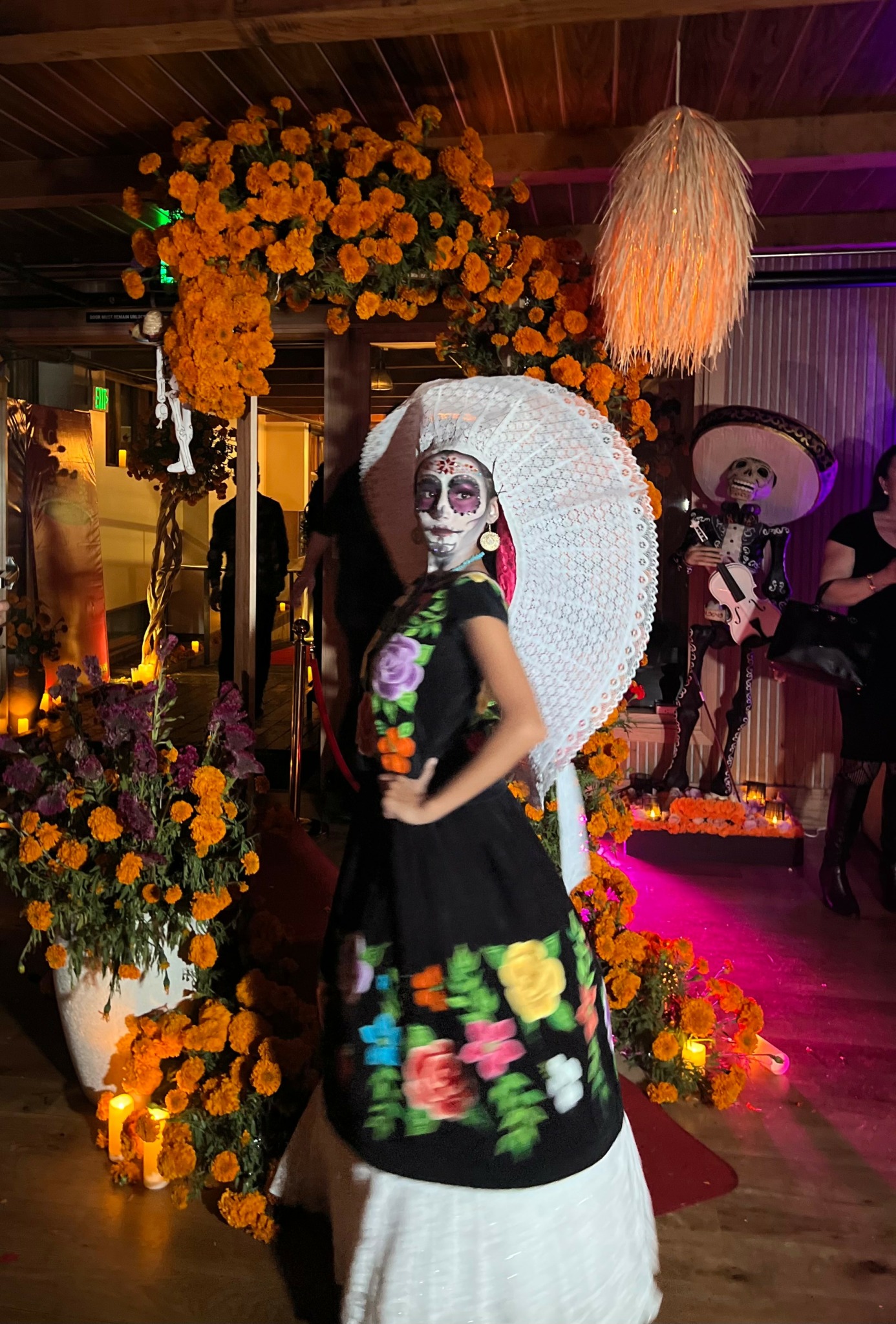 Person dressed in colorful traditional attire with face paint, standing among orange flowers and decorations.