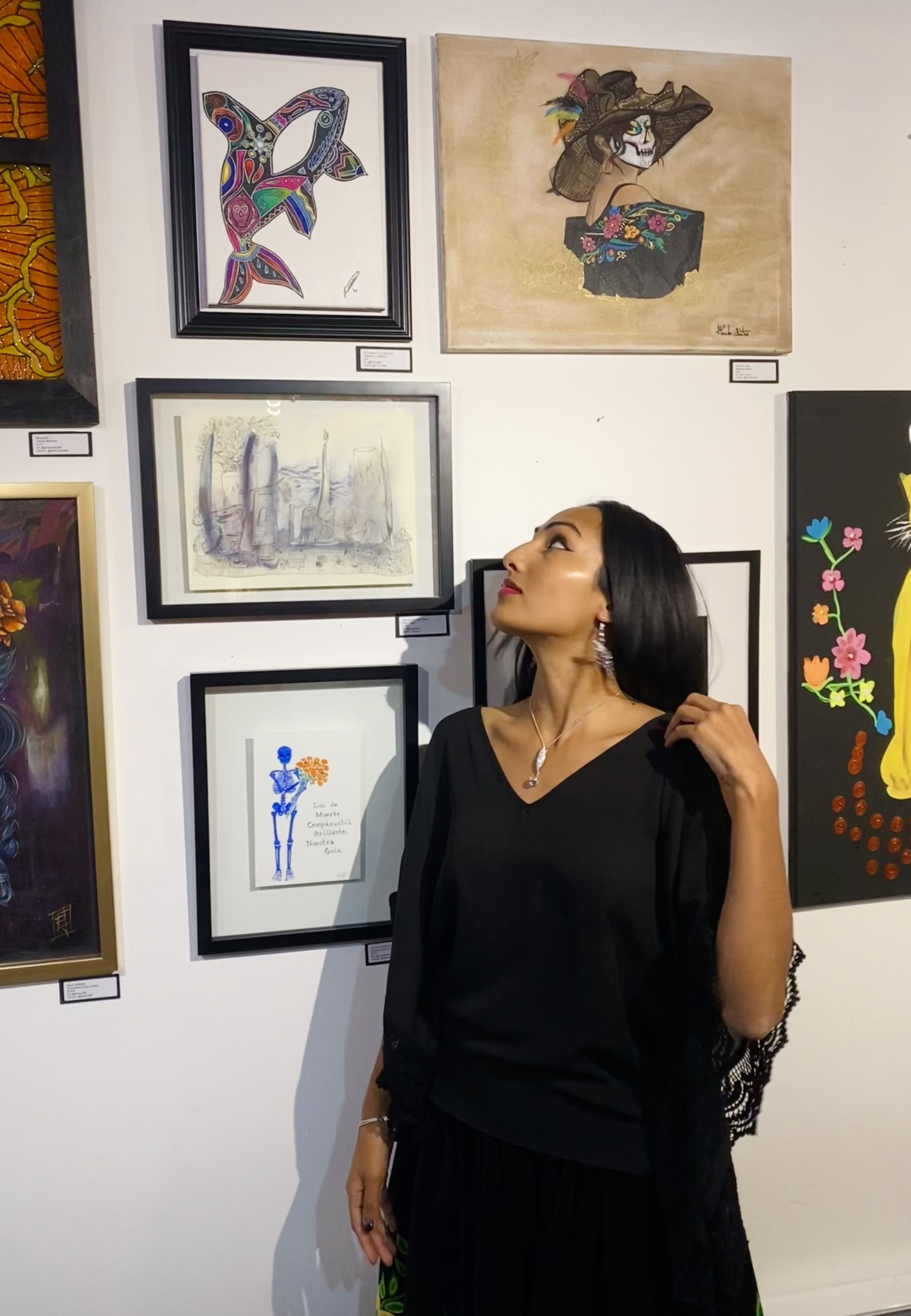 Woman with black hair wearing a black dress stands in front of a wall with framed artwork, looking to the side.