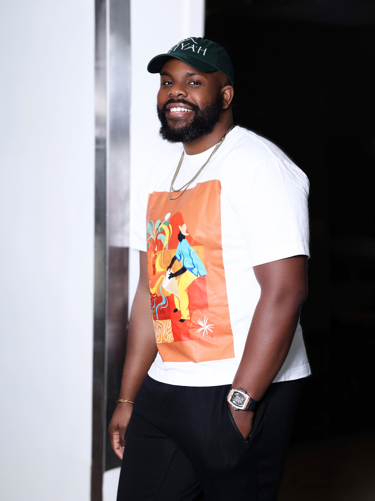 Smiling man with beard wearing a cap, white t-shirt with colorful graphic, and black pants, standing indoors.