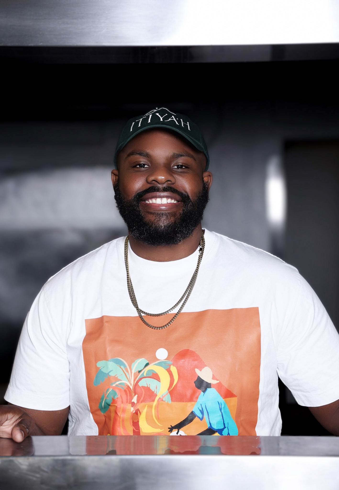 Smiling man with a beard wearing a black cap, white t-shirt with colorful graphic, and gold chains.