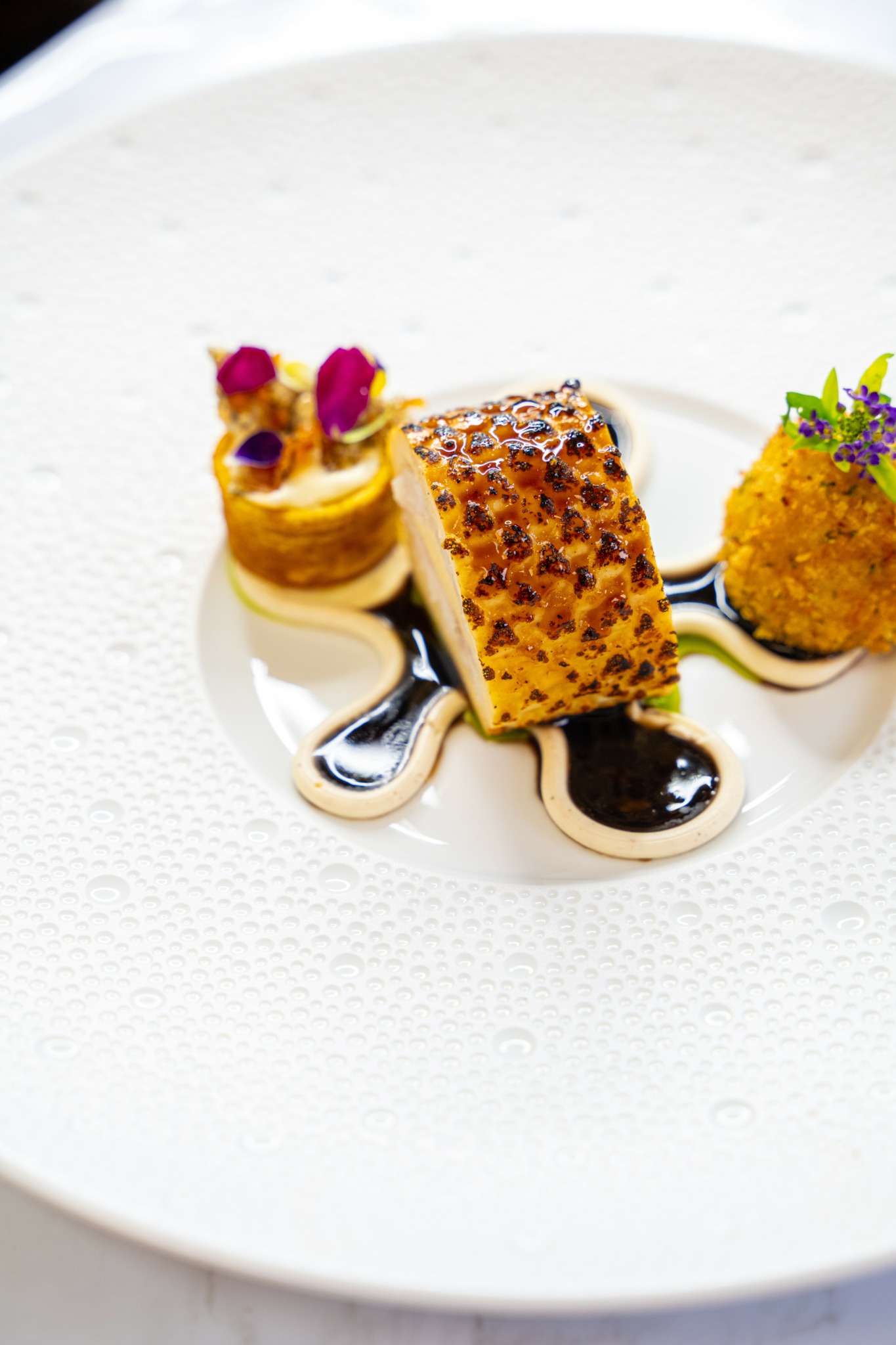 Dessert plate with a layered cake, a round pastry, and garnishes, drizzled with sauce on a white textured plate.