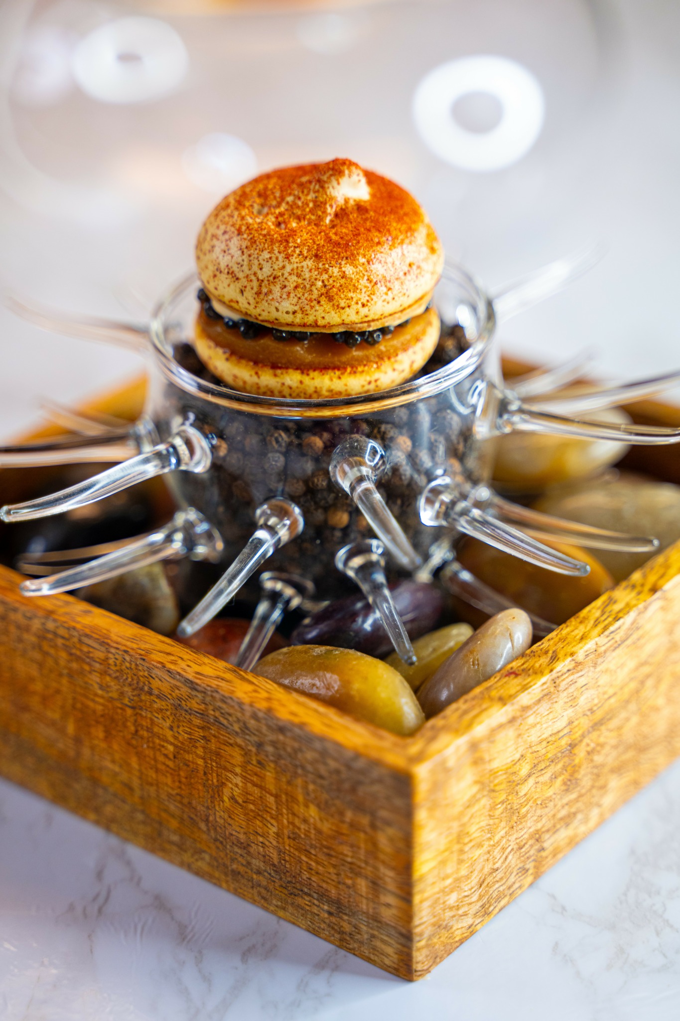 A small bread roll on top of a glass container with metal spikes, inside a wooden box with stones.