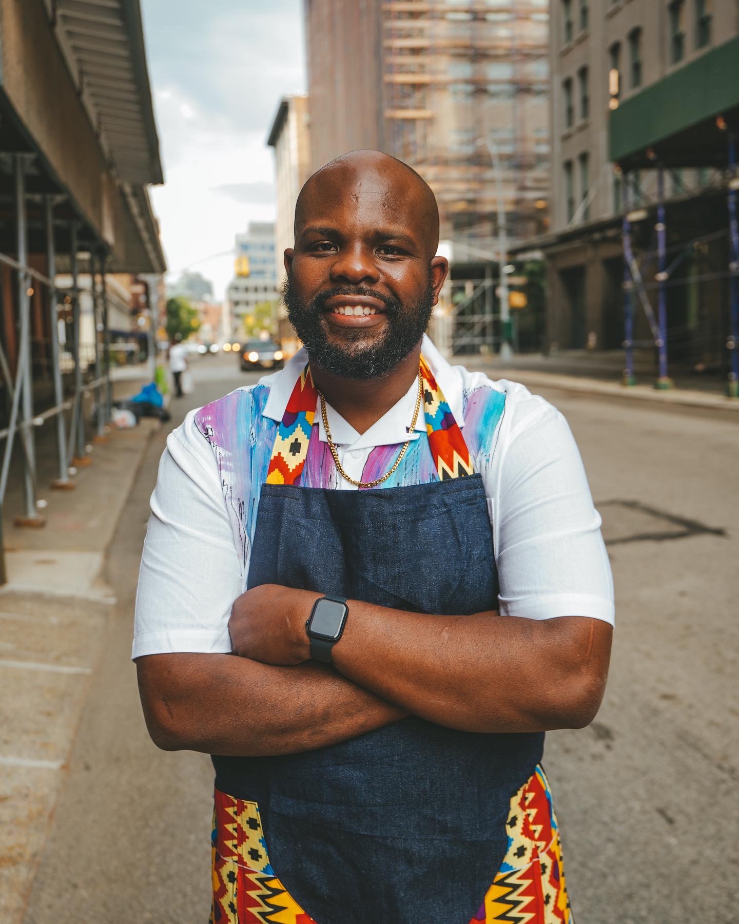 Smiling man with a beard wearing a colorful apron and white shirt, standing on city street with buildings.