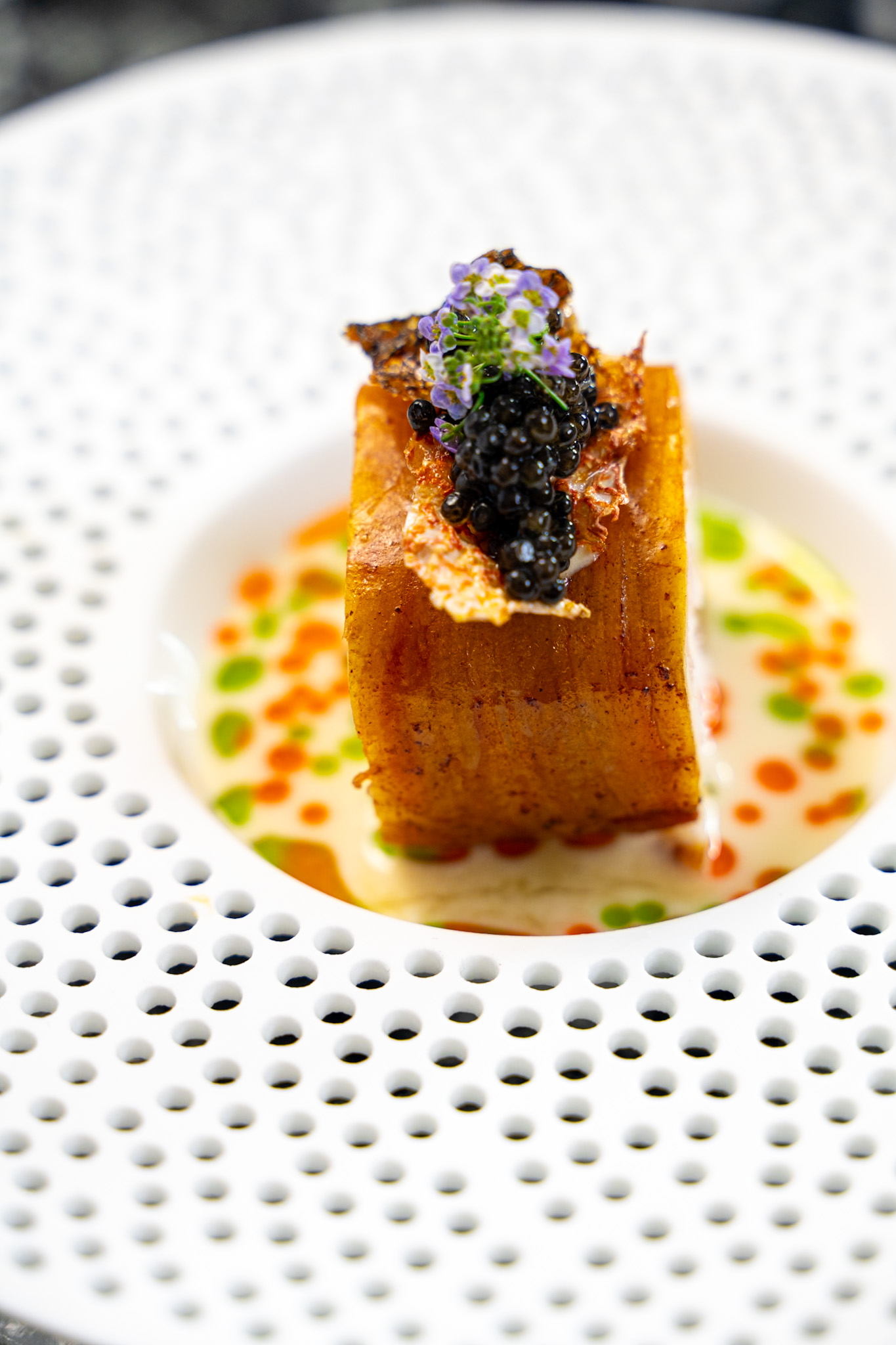 A plated dessert with a rectangular cake topped with purple flowers and blackberries, garnished with colorful sauce dots.
