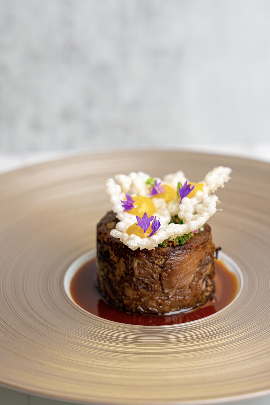 A dessert topped with white and purple edible flowers on a round plate with sauce.