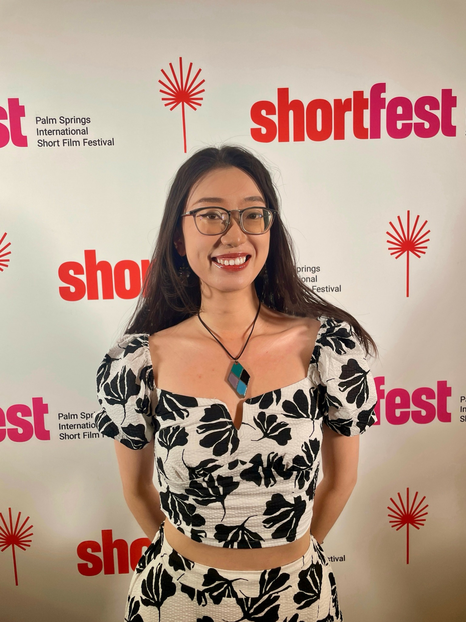 Woman with glasses smiling, wearing a black and white floral dress, standing in front of a backdrop with 'shortfest' logos.