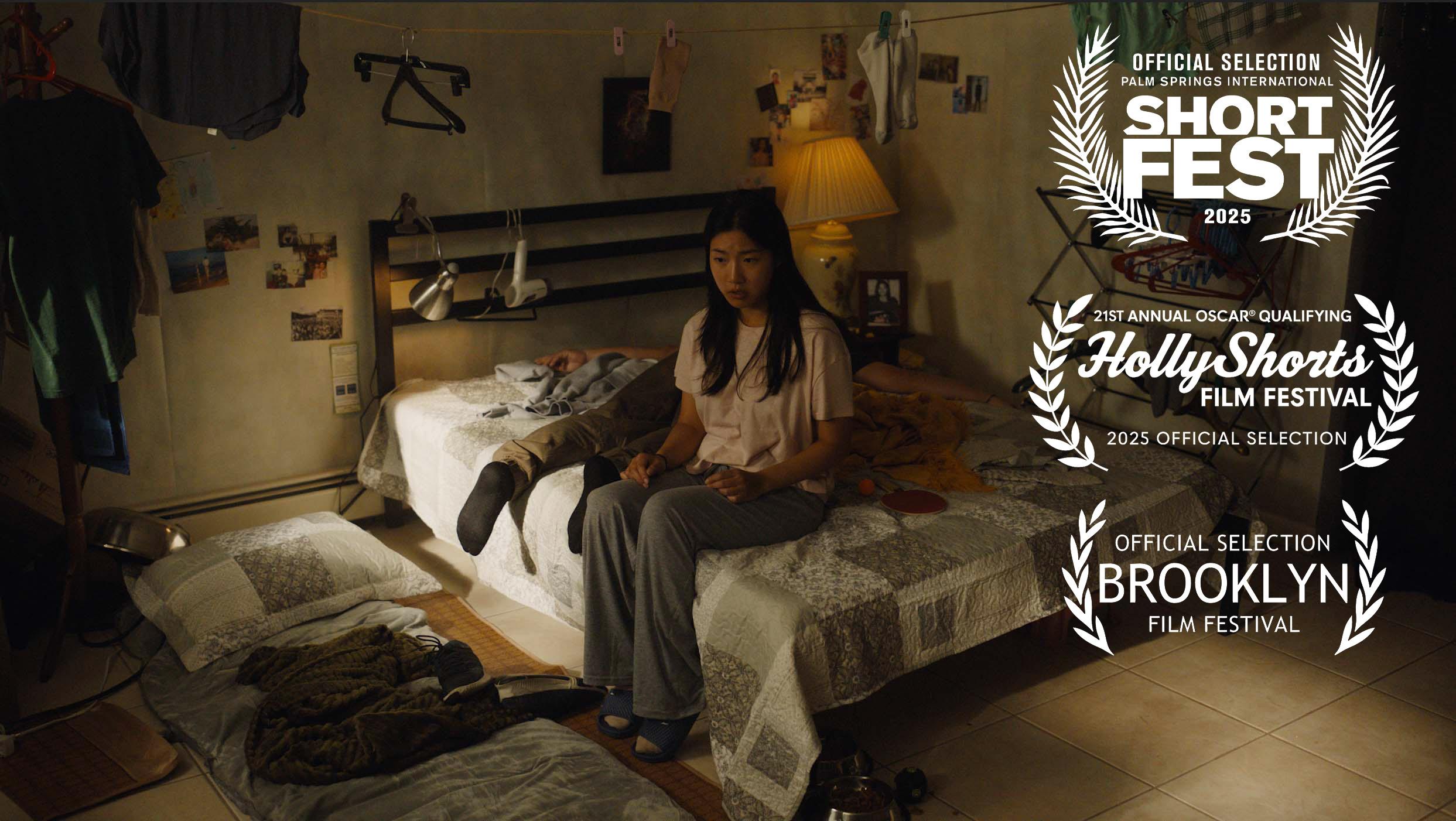 A woman sits on a bed in a dimly lit bedroom with a lamp, photos, and clothes around.
