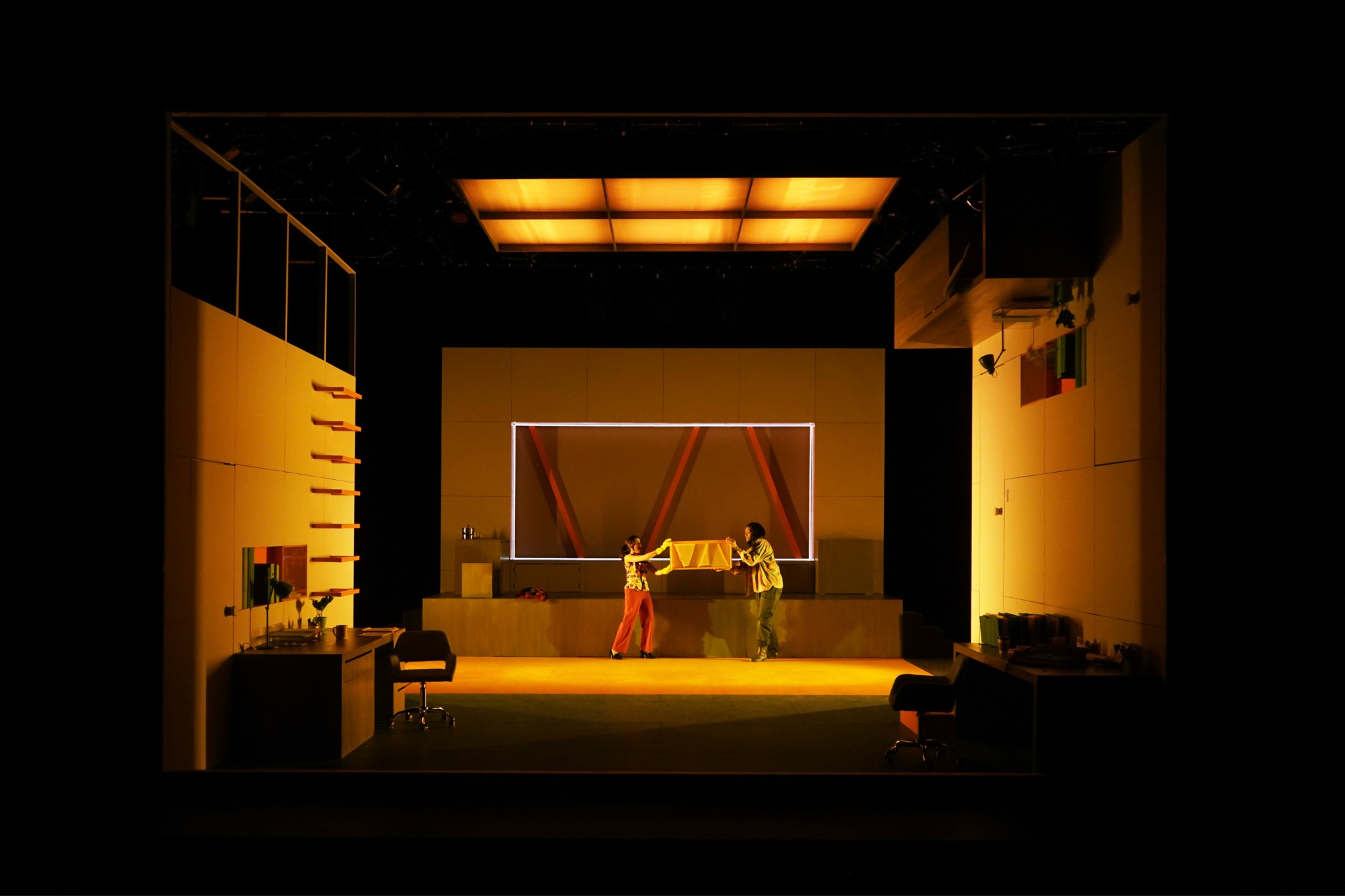 Stage with two people, one in a red dress, performing with a table and props, illuminated by warm lighting.