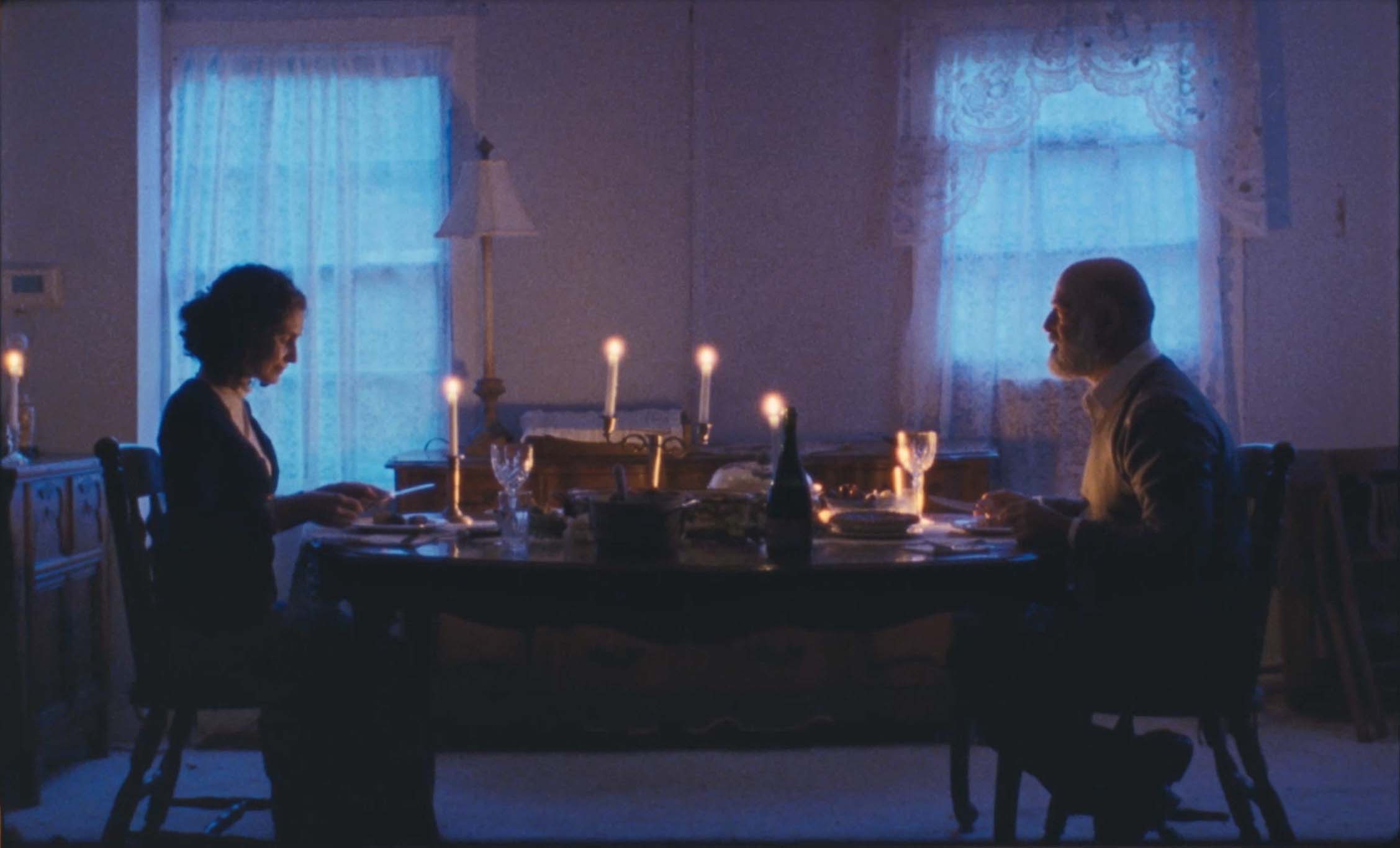 Two people sit at a dining table with lit candles, in a dimly lit room with two windows and curtains.