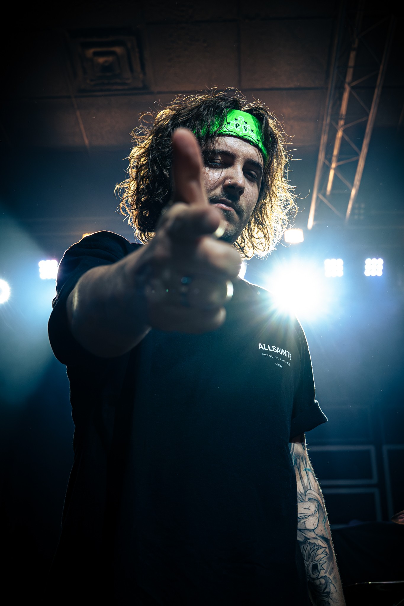 Person with curly hair wearing a green cap, pointing towards the camera, illuminated by bright stage lights.