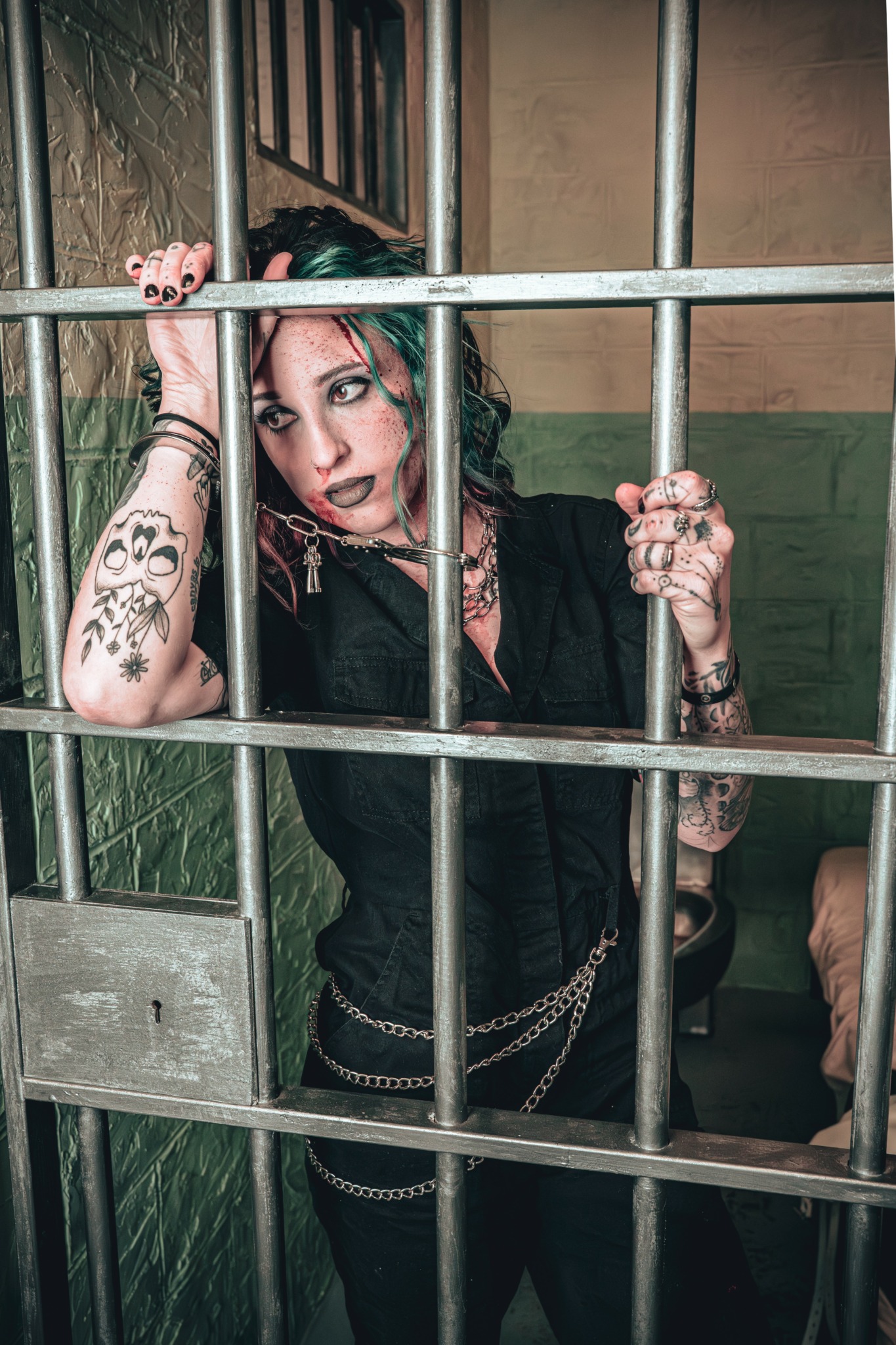 Young woman with tattoos and dark clothing behind bars, holding onto the bars with both hands, in a room with green walls.